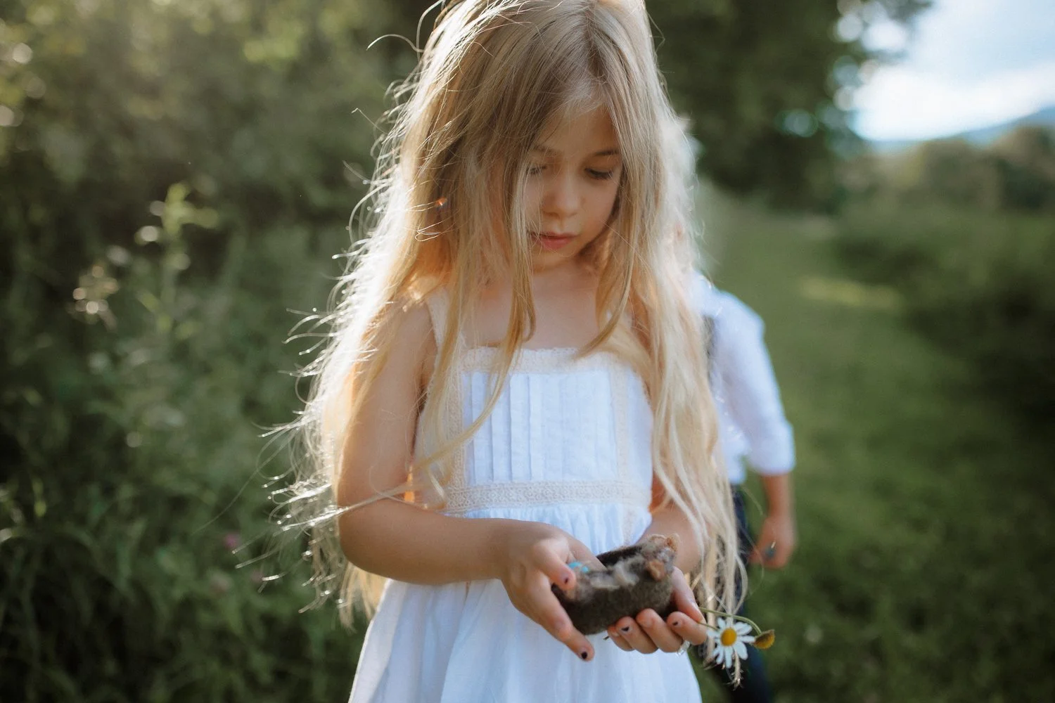 A young girl with long blonde hair in a white dress holding a small animal, possibly a hamster or mouse, in her hands, outdoors in a green, wooded area during daylight.
