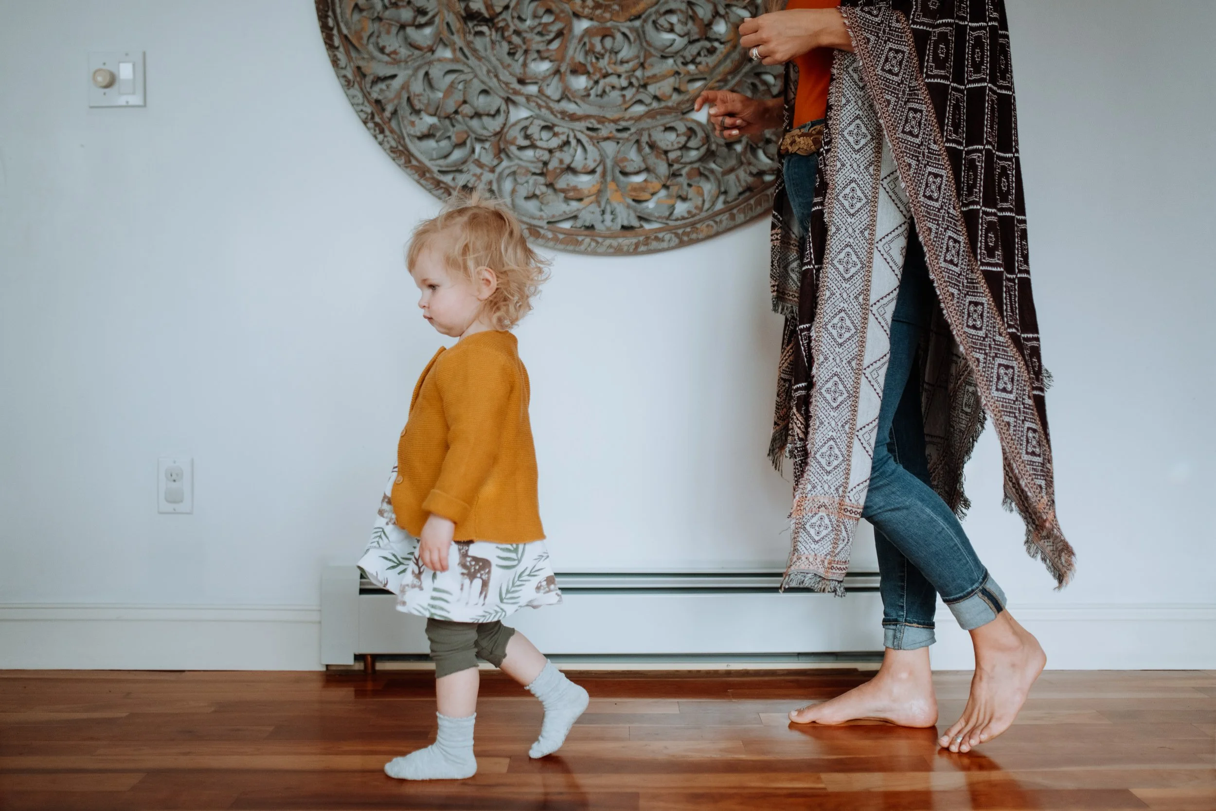 Toddler walking across a wood floor with her mother visible behind her in a patterned shawl