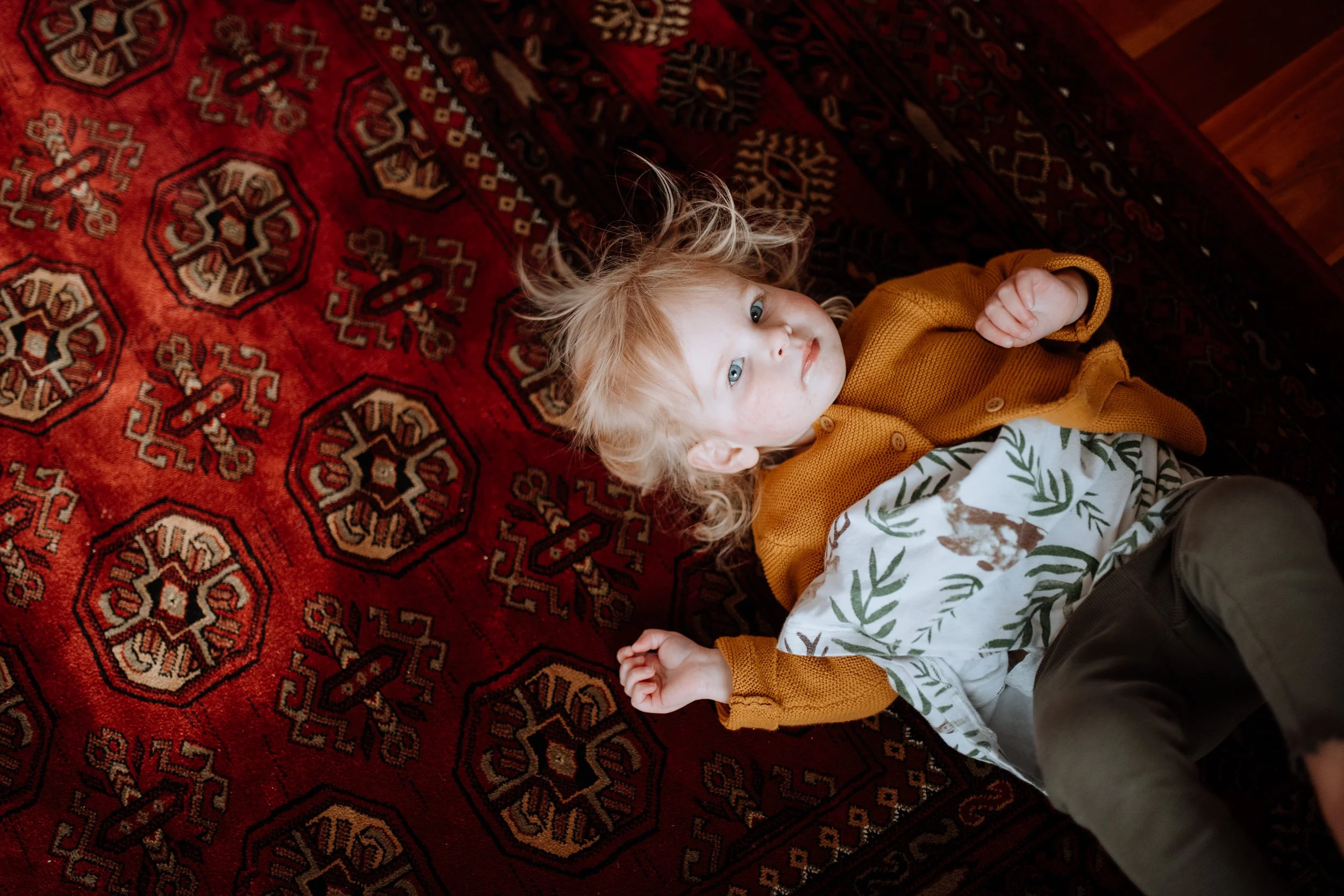 A young child with curly blonde hair and blue eyes lying on a patterned red rug, wearing a mustard yellow cardigan, a white shirt with green foliage and animal print, and olive green pants.