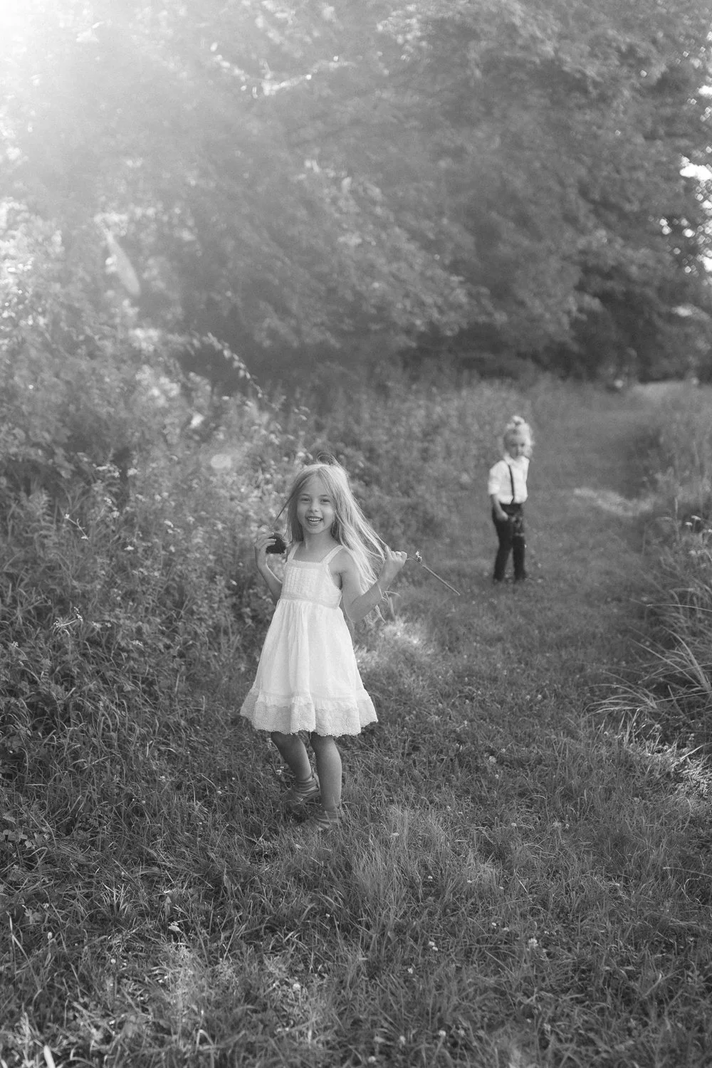 Smiling young girl in a white dress walking along a grassy trail holding a stick, with a young boy in suspenders and a white shirt in the background in a natural outdoor setting with trees.