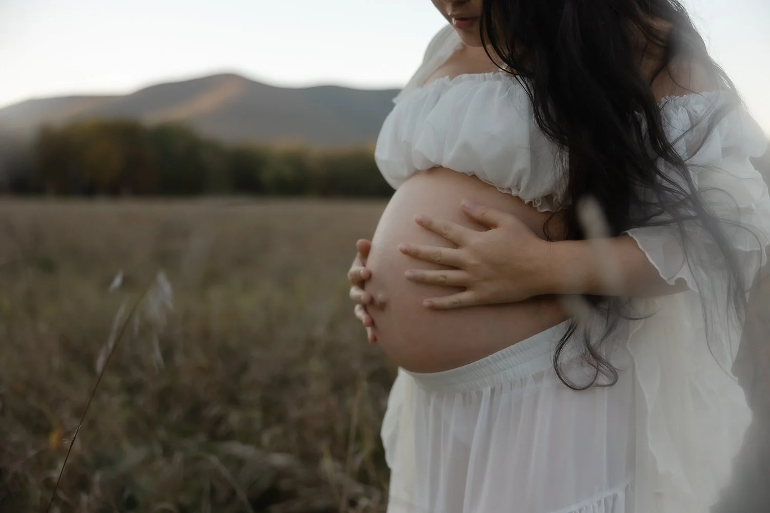 Pregnant woman stands in a field with her hands on her belly and long black hair flowing.