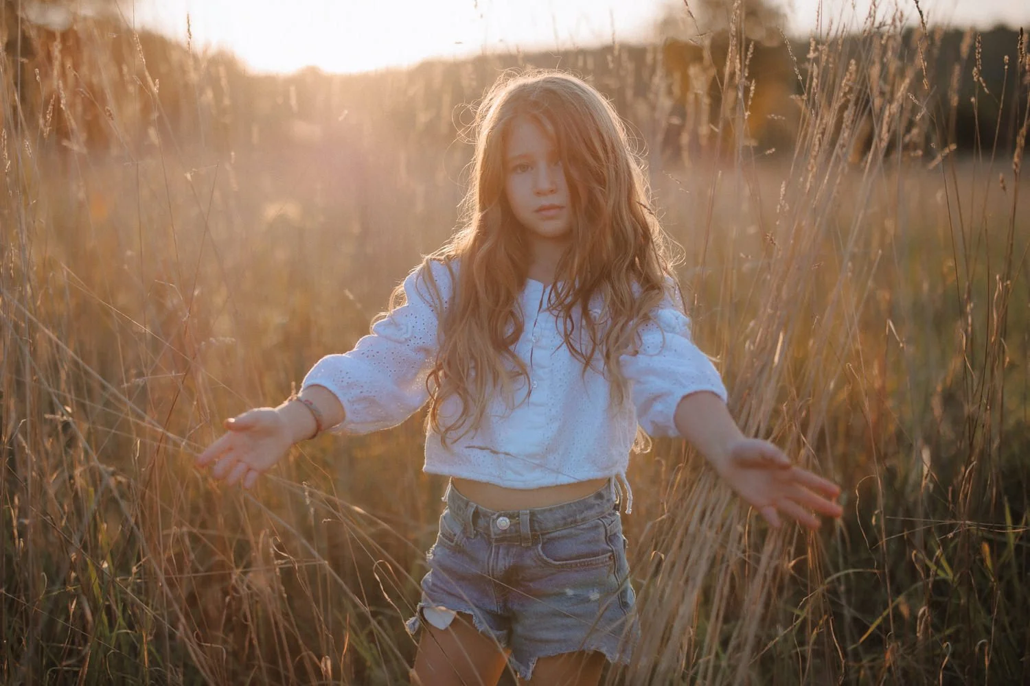A young girl with long, wavy red hair and a white long-sleeve shirt stands in a wheat field during sunset, with arms outstretched and a serious expression.