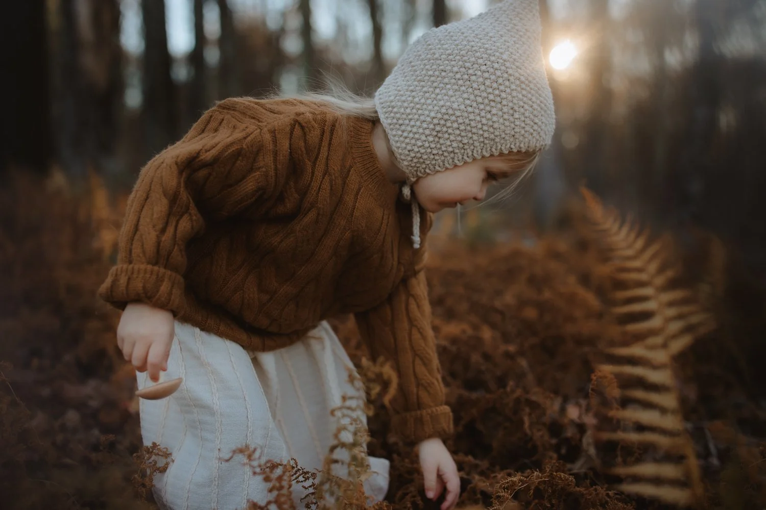 Young girl with blonde hair wearing a beige knit hat and brown sweater, leaning over to touch the soil in a forest during autumn.