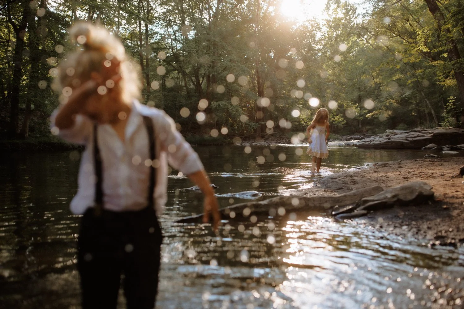 Two young girls in a forest stream, one in the foreground with blurred details, the other in the background wearing a white dress, surrounded by trees and sunlight.