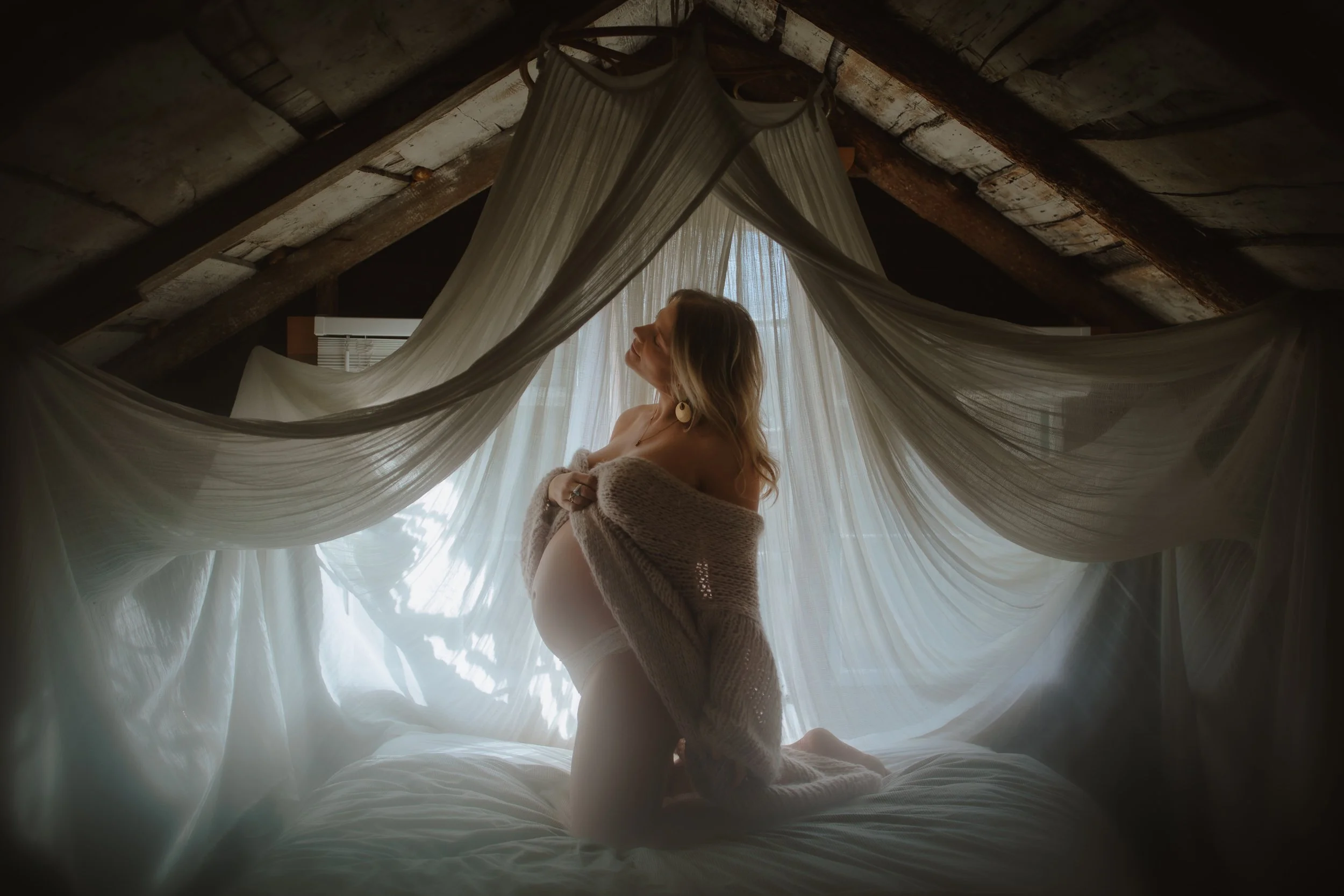 Dreamy portrait of a pregnant woman kneeling beneath a sheer curtain canopy in an attic