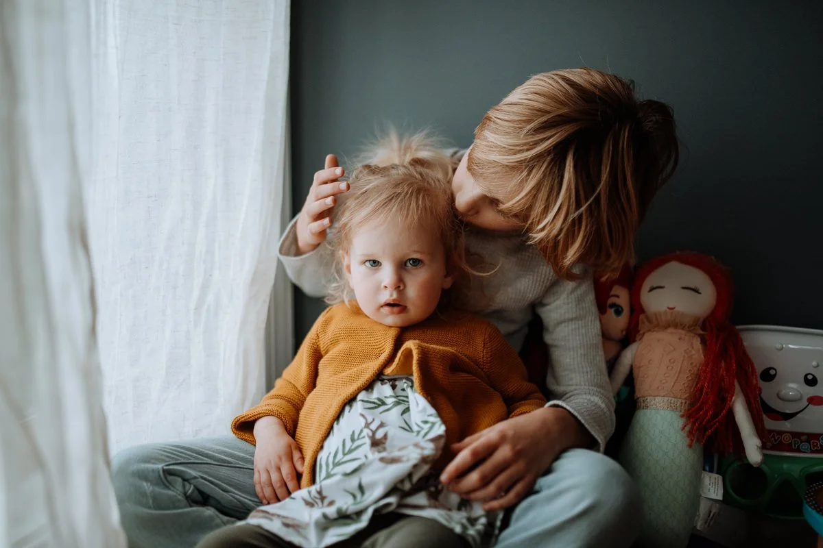 A woman with light brown hair kissing a young girl on the head, while the girl sits on her lap looking at the camera with a serious expression. The girl has blond hair and is wearing an orange cardigan. The setting appears to be indoors near a window