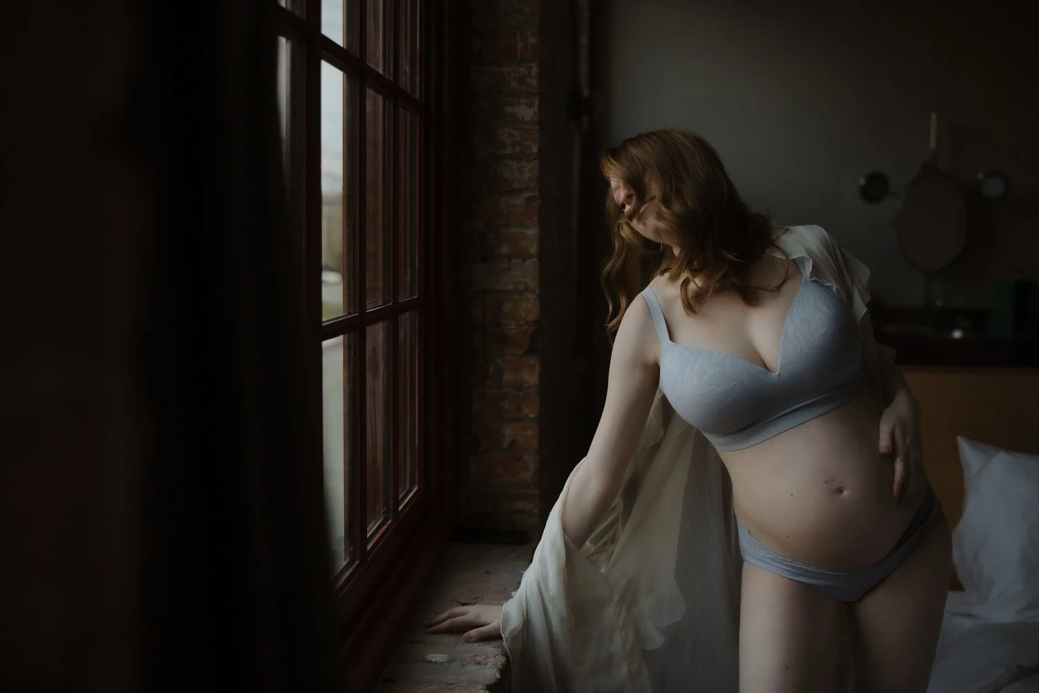 Pregnant woman in a blue bra looking out a window in an industrial brick space