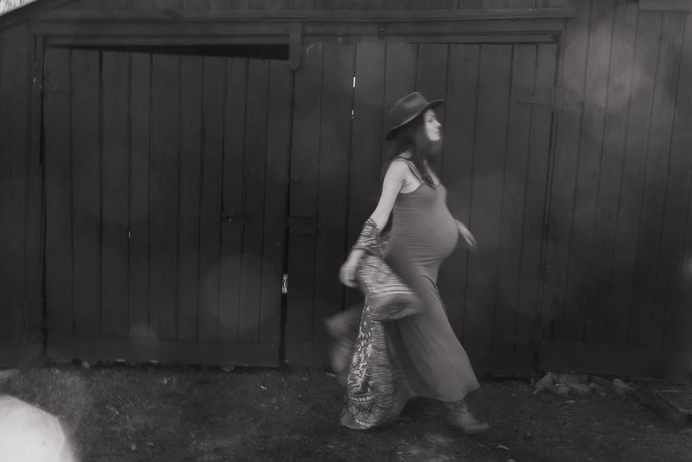 Black and white portrait of a pregnant woman in a wide-brimmed hat walking past a wooden fence