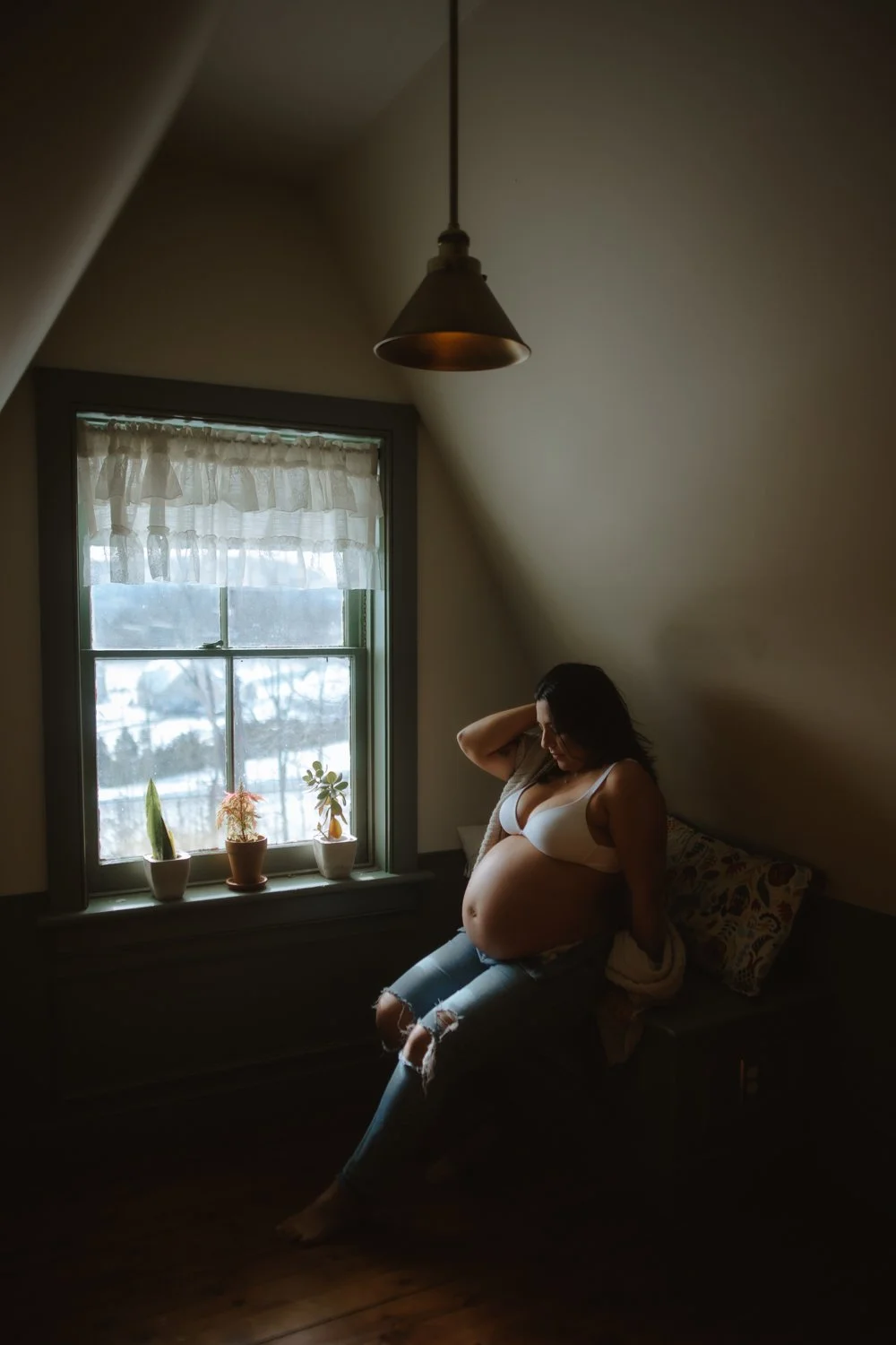 Pregnant woman sitting on a window bench in soft natural light, hand in her hair, potted plants on the sill behind her.