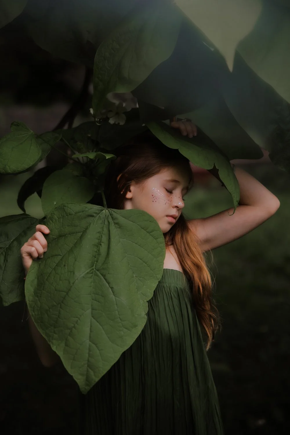 A young woman with long reddish hair and glitter on her face is surrounded by large green leaves, with her eyes closed and one arm raised above her head, wearing a dark green strapless dress.