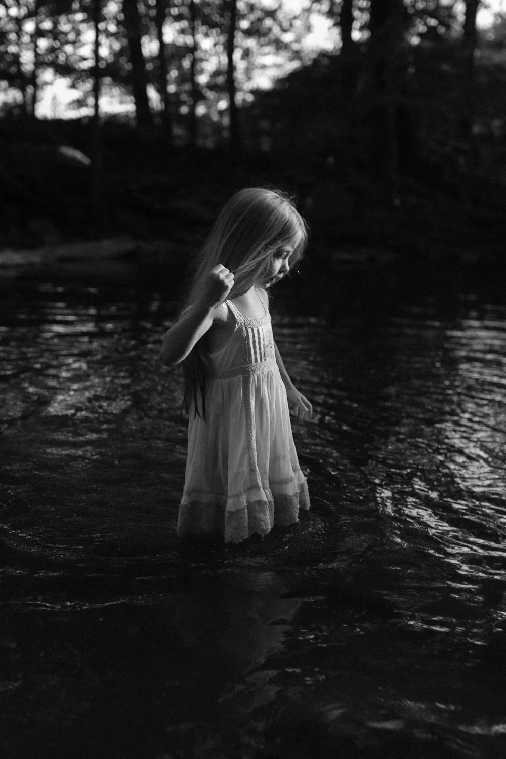 A young girl in a dress standing in a body of water, with trees and a sunset or sunrise in the background, captured in black and white.
