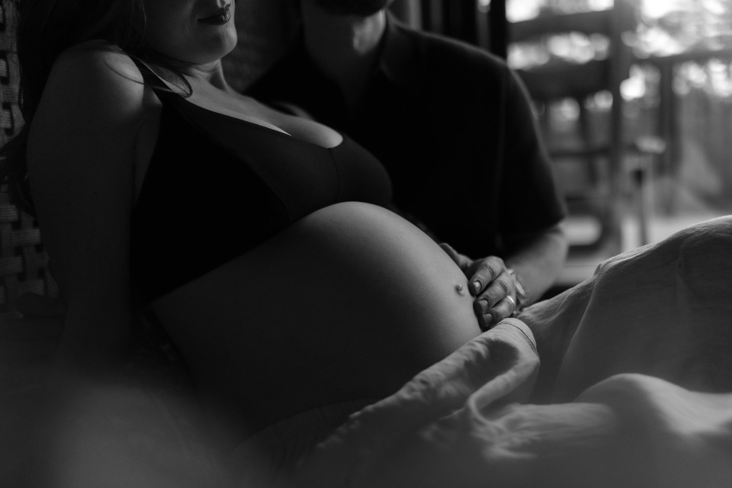 A pregnant woman lying on a bed, shirtless, with her hand on her belly, accompanied by a partner whose face is not visible, in a dark room with natural light coming from a window.