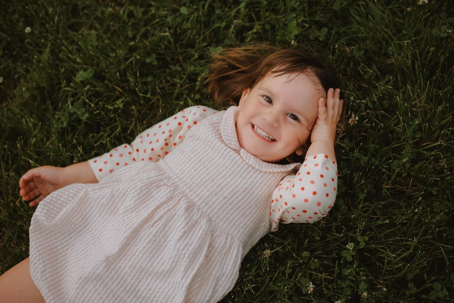 Young girl lying on grass with a happy smile, wearing a cream-colored dress with red polka dots, and resting her head on her hand.