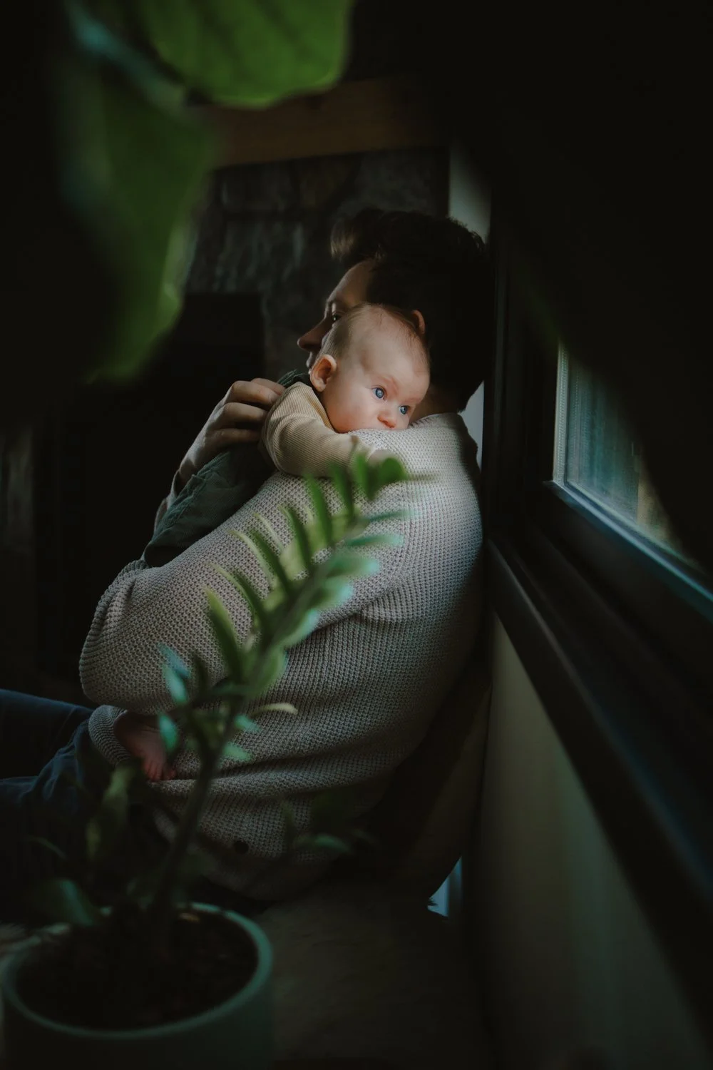 Baby peeking over father's shoulder in soft window light, captured by a baby photographer