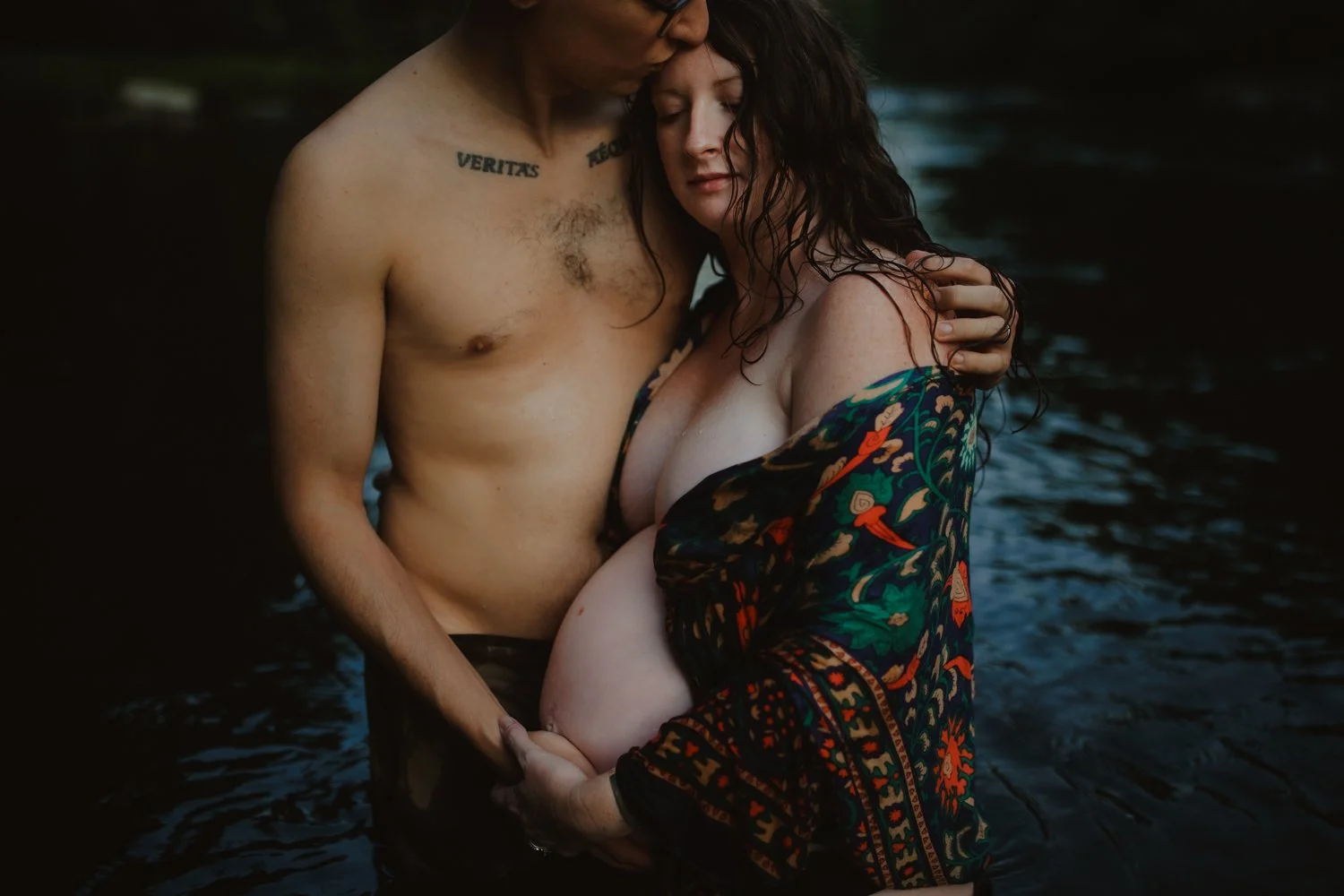 A couple standing in water, embracing each other closely. The man is shirtless, and the woman is partially wrapped in a colorful, patterned shawl or robe. They are holding hands, with the woman's hand on her pregnant belly, and they are gently touching foreheads with their eyes closed.
