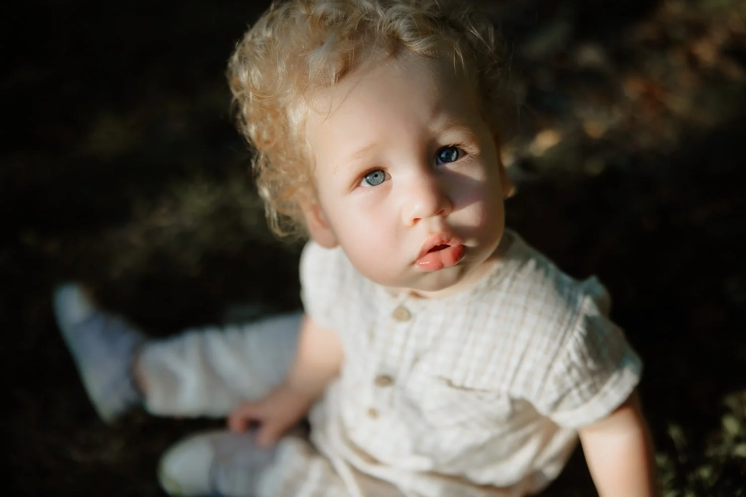 Young child with blonde curly hair and blue eyes looking up outdoors, sitting on dark ground, wearing a light-colored outfit.