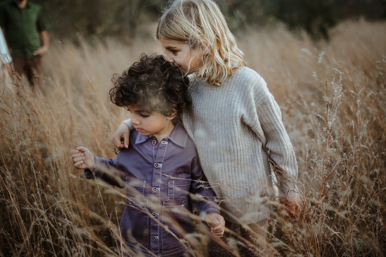 A woman and a young boy walking through a field of tall, dry grass. The woman has blonde hair and is wearing a light gray sweater, and she is gently leaning her head on the boy's head. The boy has dark, curly hair and is wearing a dark purple jacket,
