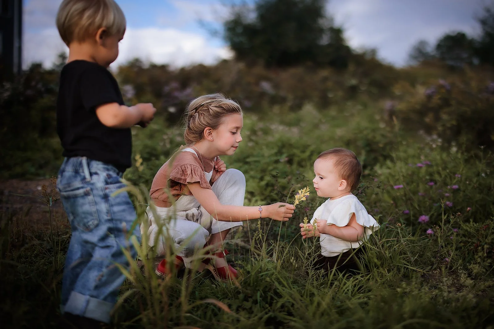 Three young siblings in a wildflower field, an older girl handing a flower to a toddler while another child looks on.