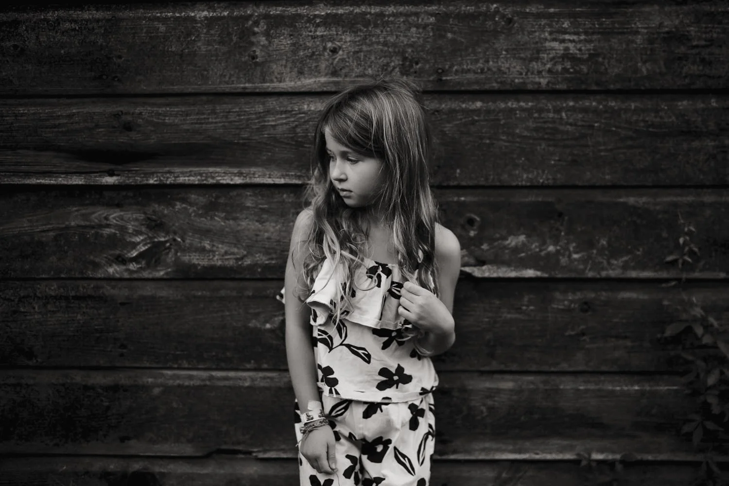 A young girl with long, wavy hair stands against a dark wooden background. She is looking down and to the side, wearing a floral patterned outfit.