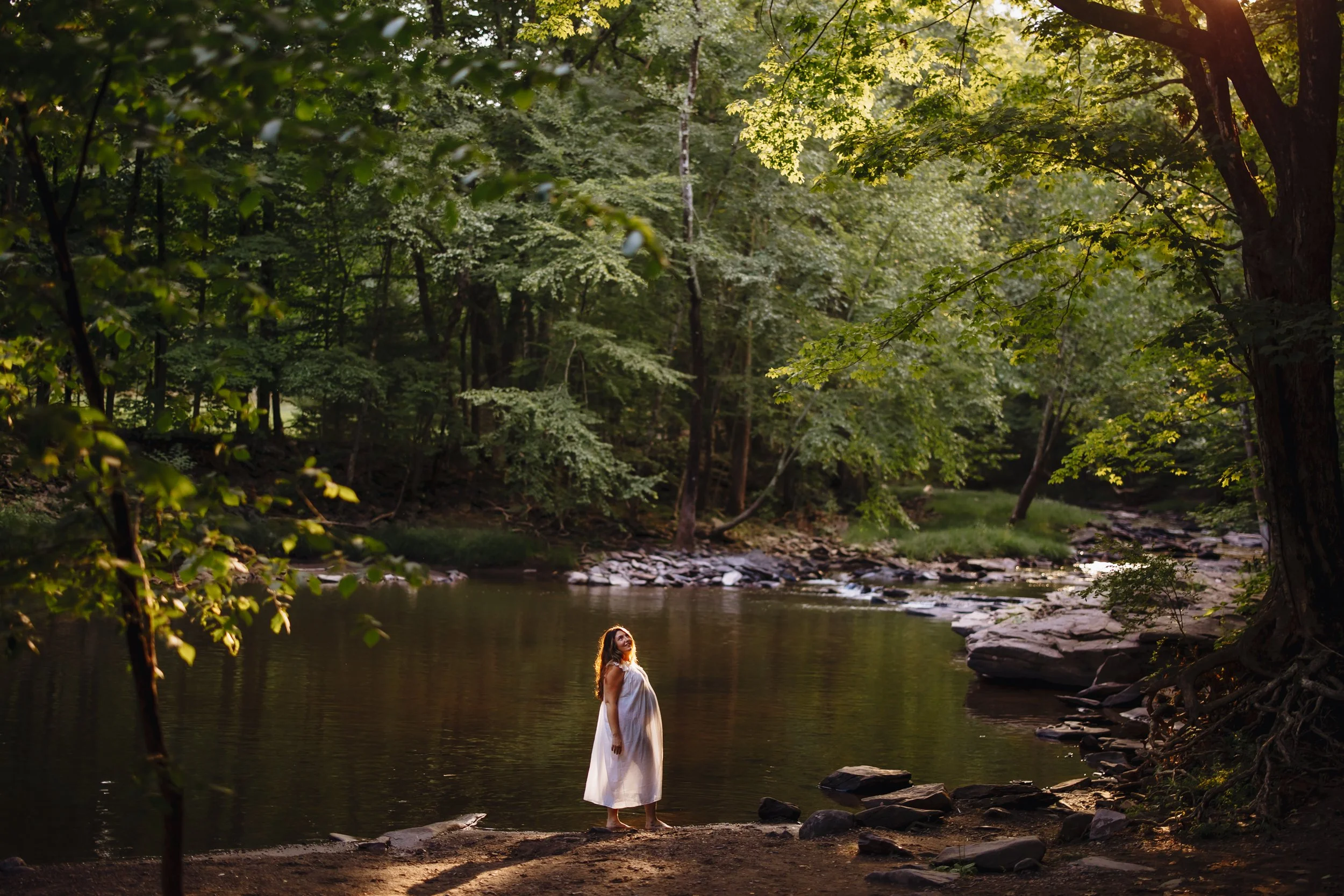 Romantic portrait of a pregnant woman in a white dress standing in a wooded creek
