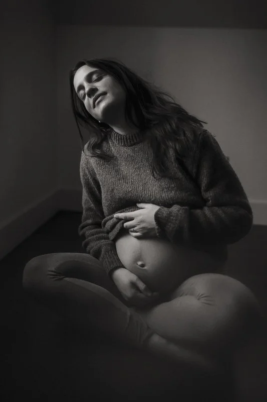 Black and white portrait of a pregnant woman cradling her belly in a knit sweater