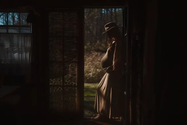 Pregnant woman in a wide-brim hat standing in a dark doorway, silhouetted against the evening light.