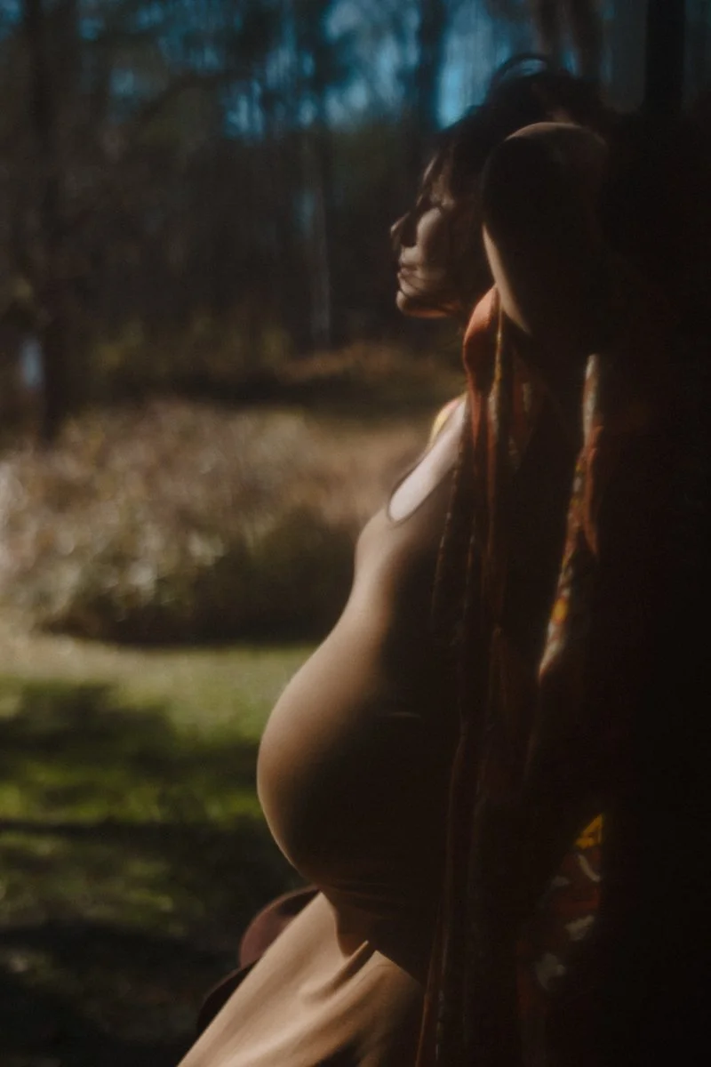 Silhouette of a pregnant woman's profile outdoors at golden hour, wrapped in a patterned shawl.