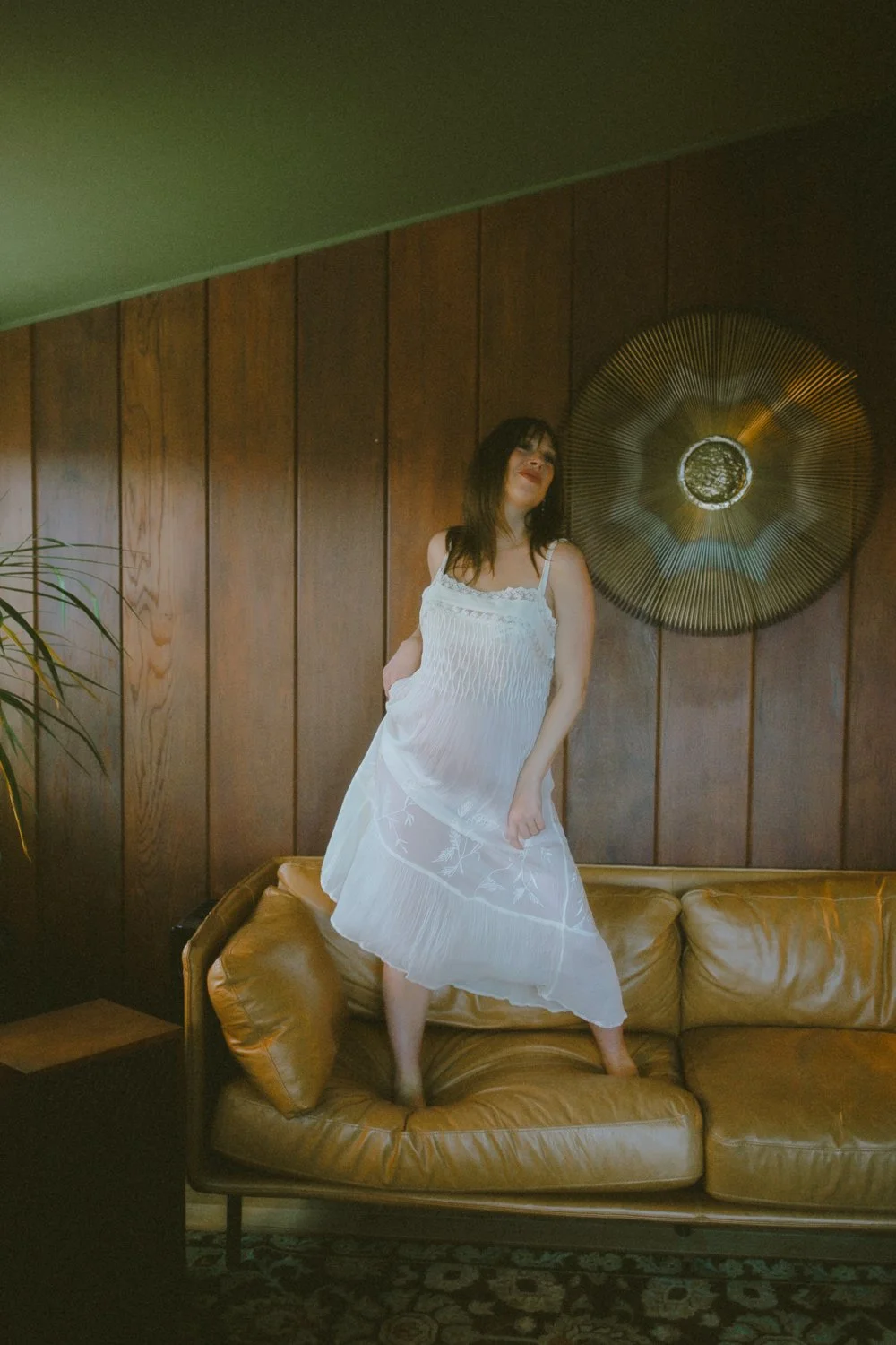 Pregnant woman in a white slip dress standing on a vintage couch in a wood-paneled room