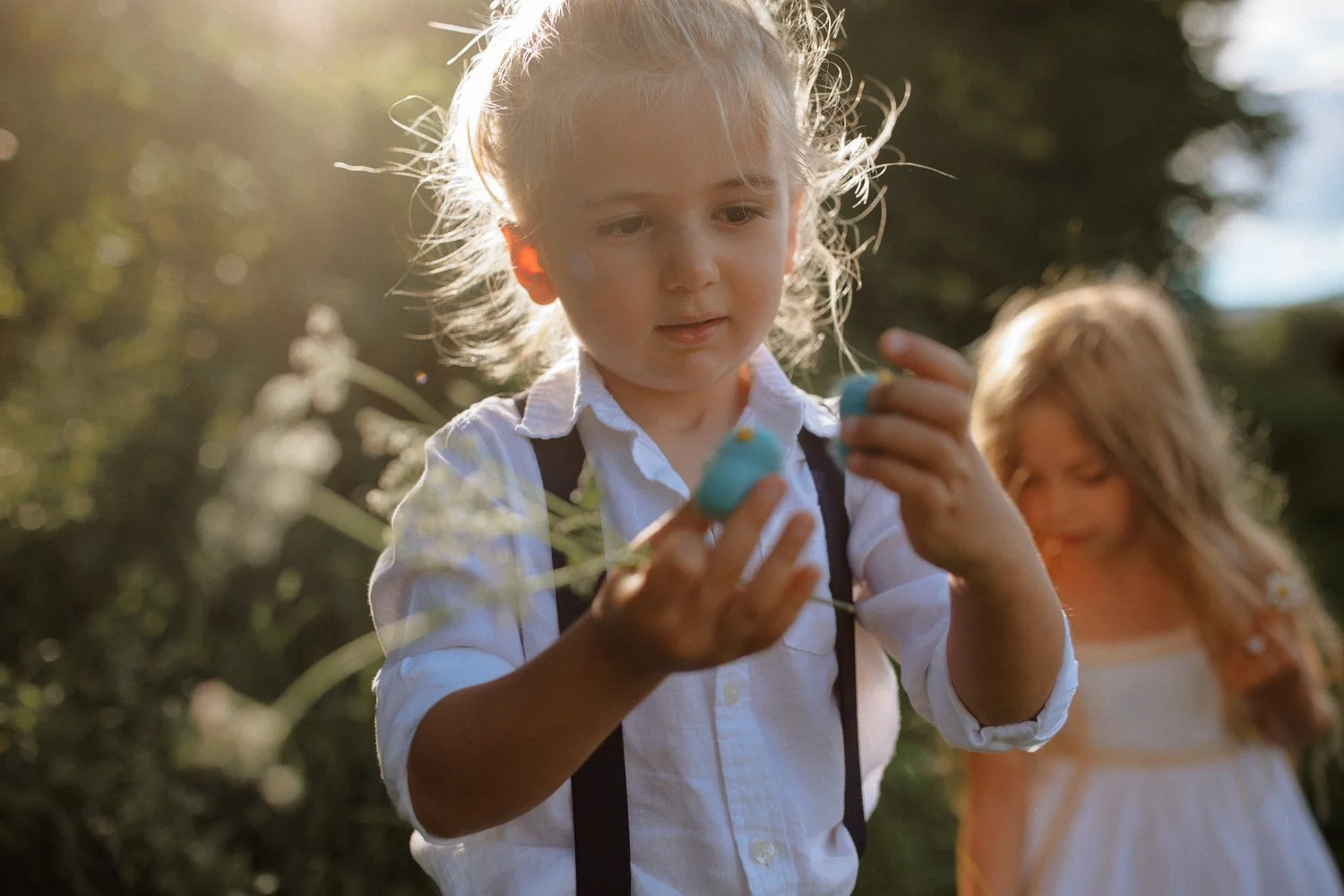 Two young girls outside in the sunlight, with one girl holding a small blue object and the other girl smiling in the background.