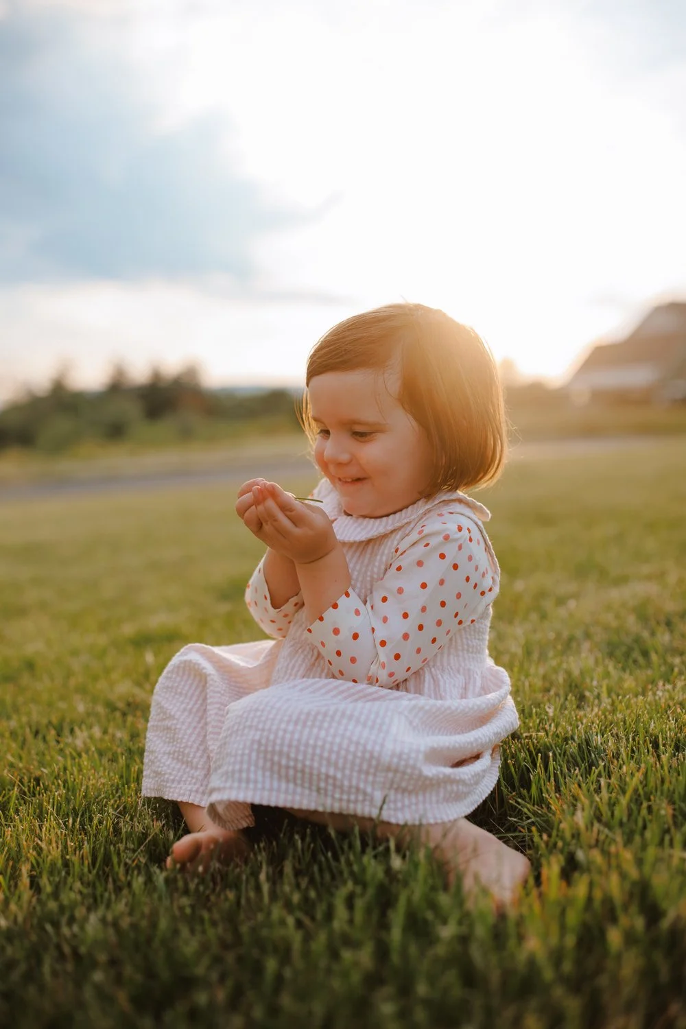 A young girl sitting on grass outdoors during sunset, wearing a pink polka-dotted dress, smiling and looking at her hands.