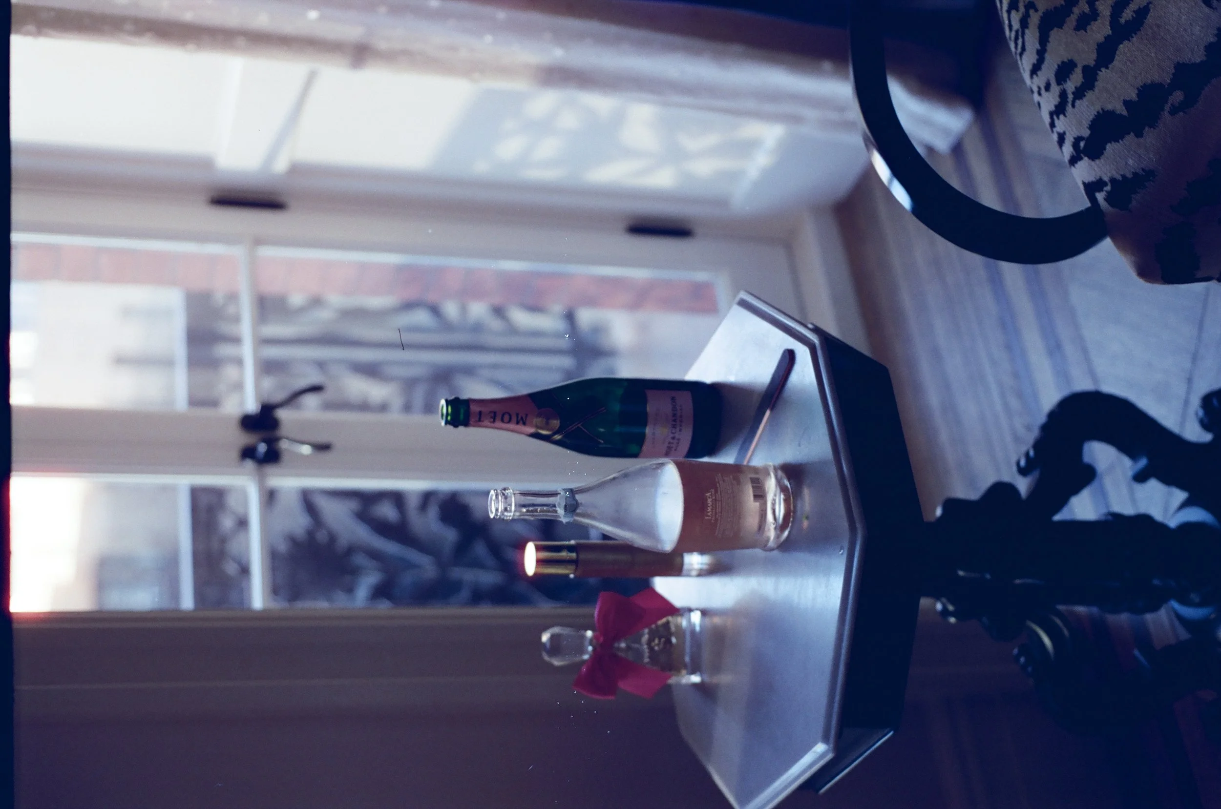 A table near a glass door with a bottle of Moët champagne, a small glass bottle with a pink bow, a conical flask, a tube with liquid, and a golden object, with a view of the outside through the door.