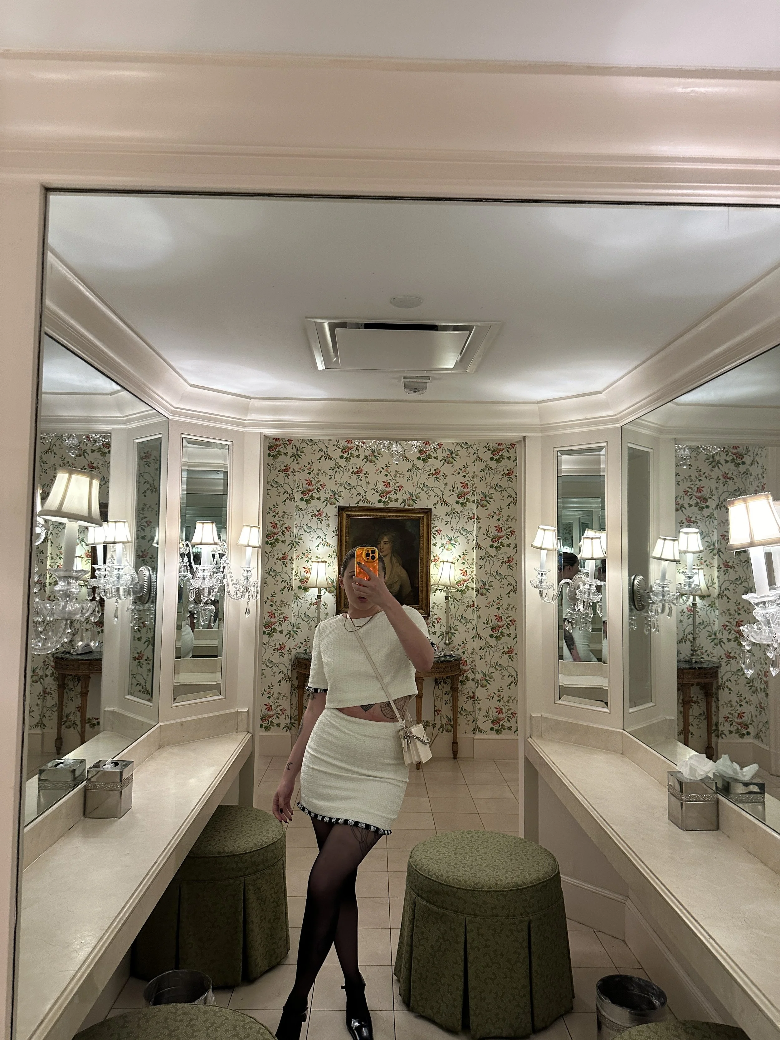A woman taking a mirror selfie in a decorated hotel bathroom with floral wallpaper, framed portrait, wall sconces, green upholstered stools, and makeup tissue box.