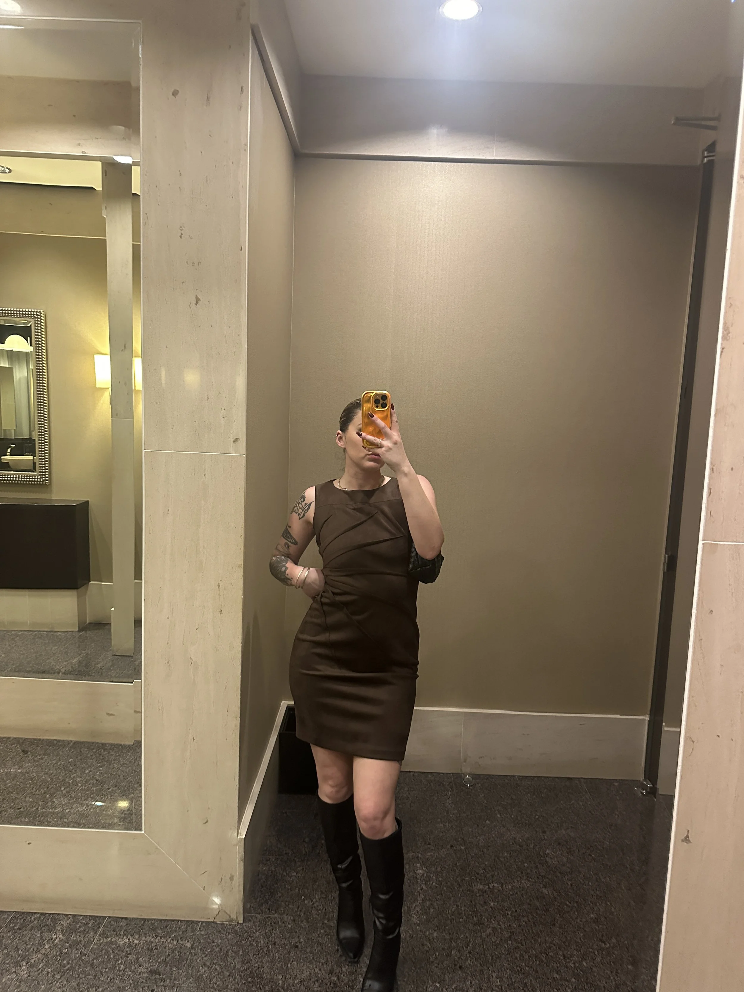 A woman taking a mirror selfie in a bathroom, wearing a sleeveless brown dress and knee-high black boots, with tattoos on her left arm.