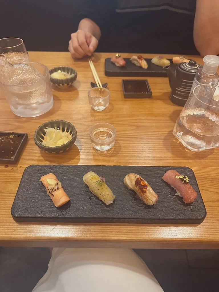 Assorted pieces of sushi on a black slate serving board with drink glasses, chopsticks, and small dishes on a wooden table.