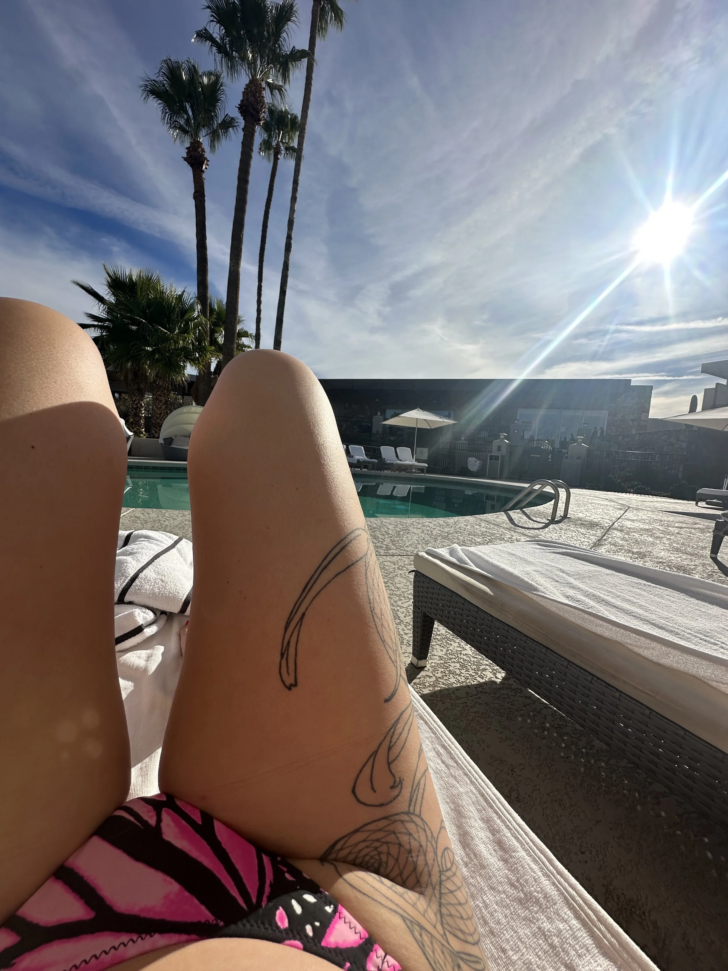 Person lounging on a poolside chair under a sunny sky with palm trees in the background.