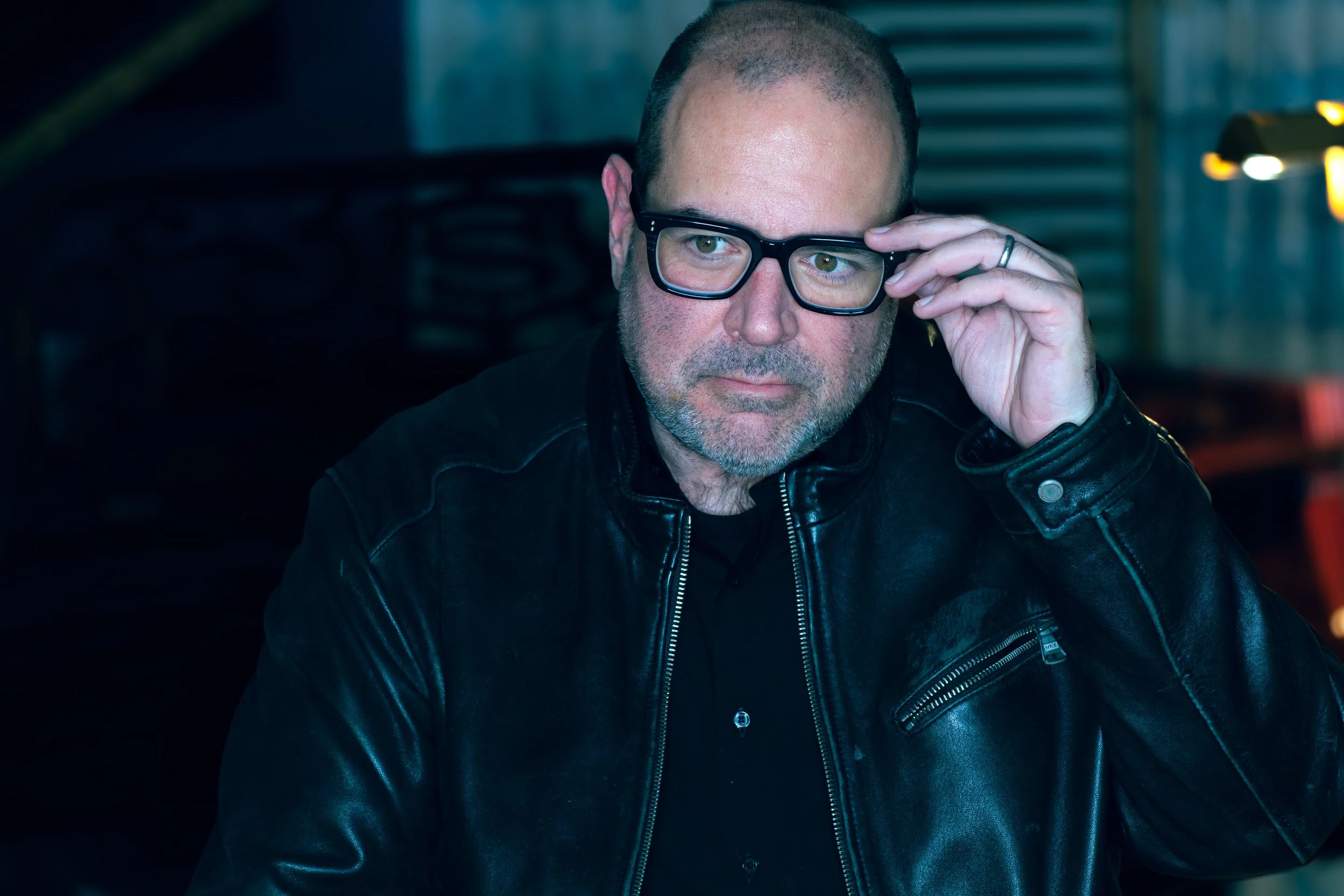 Man wearing black leather jacket and glasses adjusting his glasses indoors.