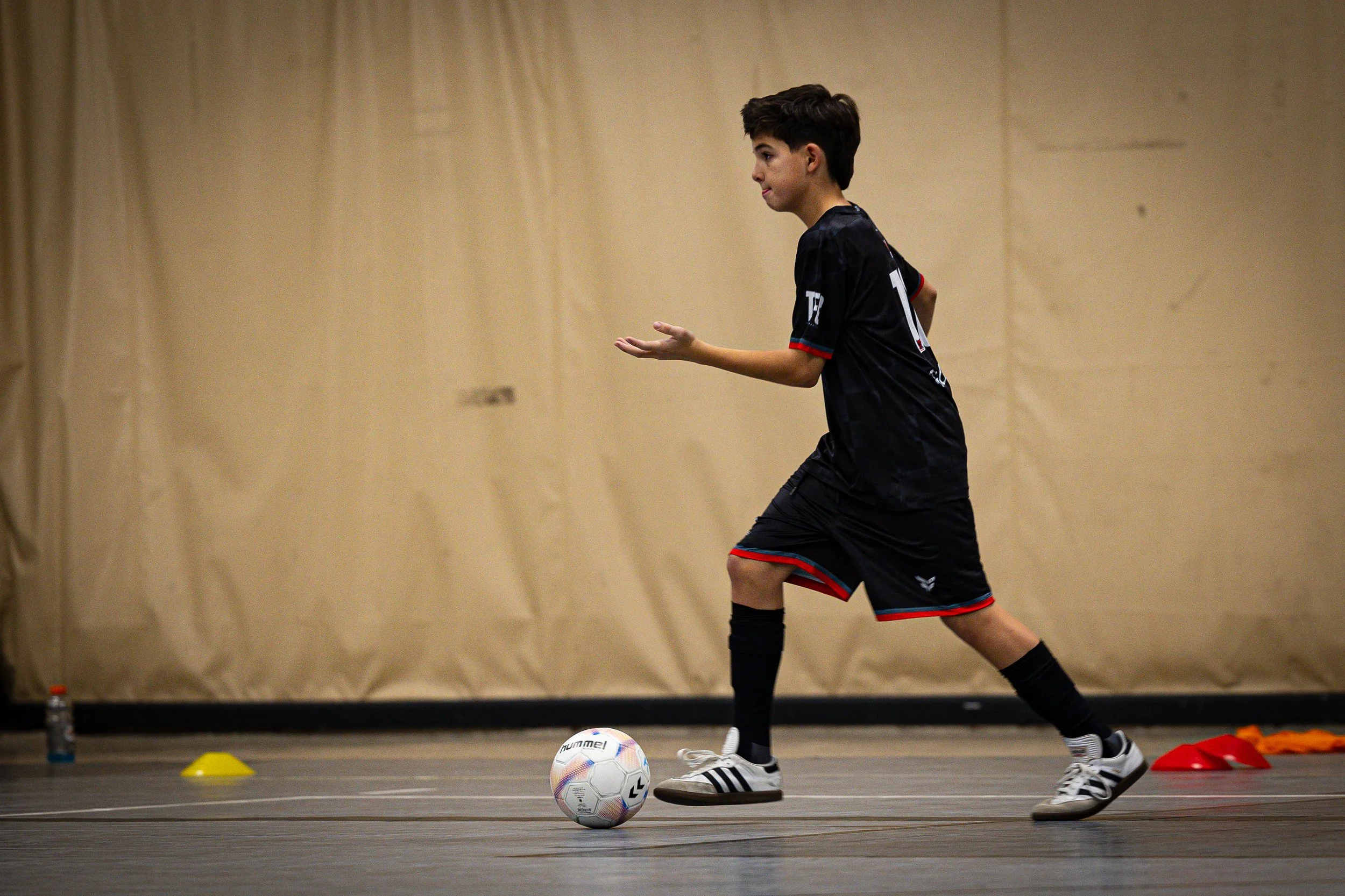 A young male athlete wearing a black sports uniform is practicing soccer indoors, dribbling around cones and a soccer ball on the floor.