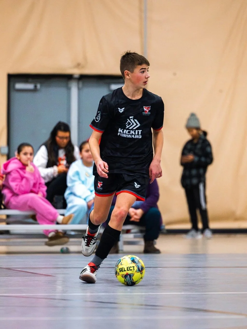 A young boy in a black soccer uniform playing indoor soccer with a yellow and blue soccer ball.