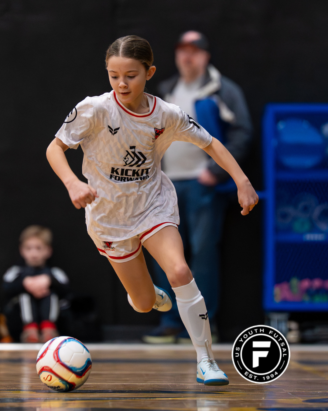 Young girl playing futsal indoors, wearing a white jersey and shorts, dribbling a soccer ball, with a person and a child sitting in the background