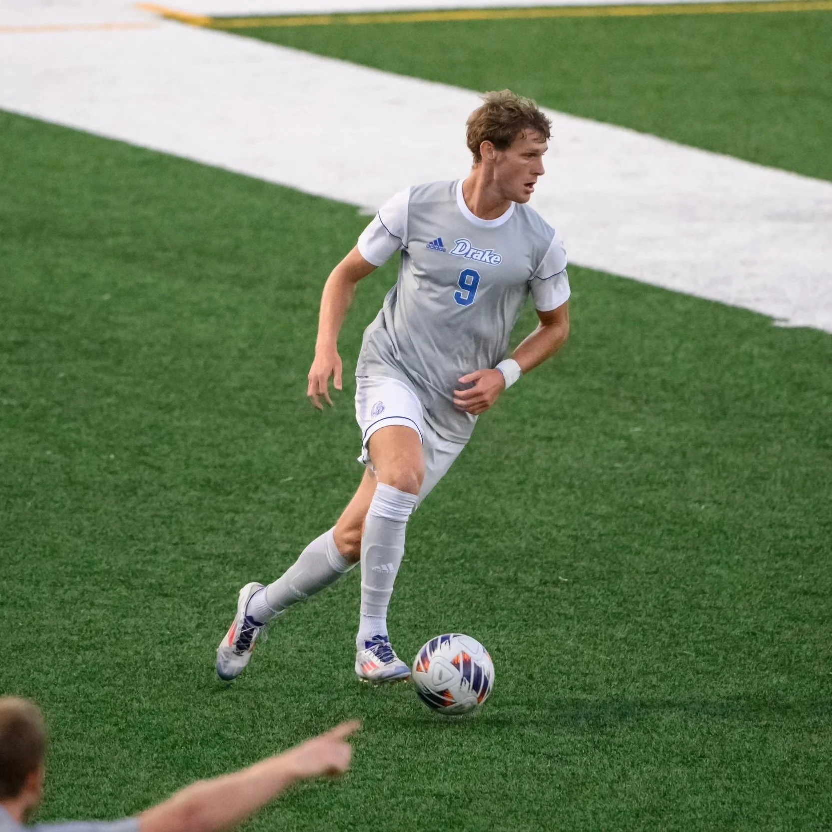 A male soccer player in gray and white uniform with the number 9 and 'Duke' on the jersey, running on a green field and about to kick a soccer ball.