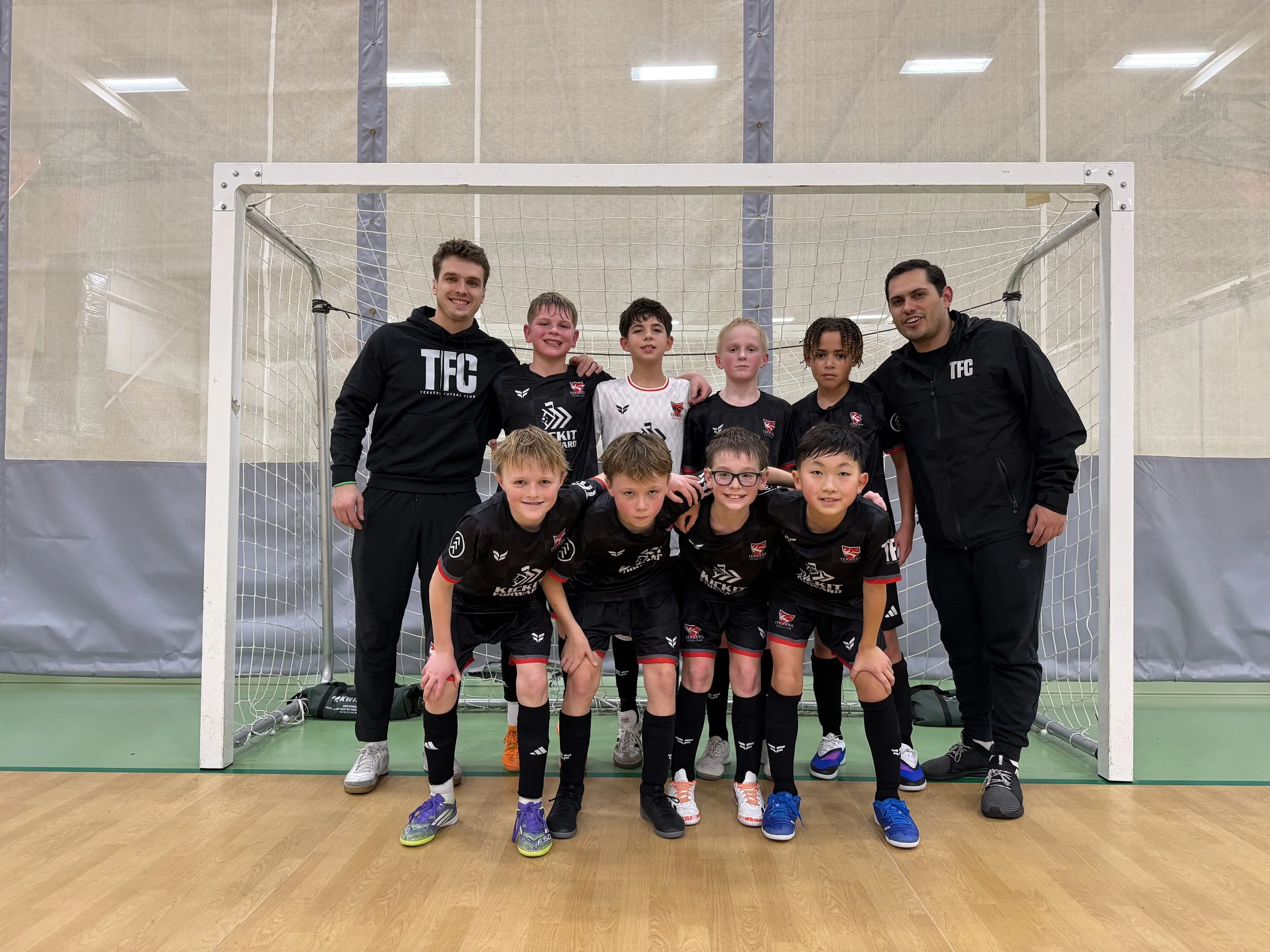 A group of young boys and two adult coaches posing for a team photo behind a soccer goal inside an indoor sports facility. The boys are wearing black soccer uniforms, and the coaches are dressed in black jackets with the logo 'TFC'.