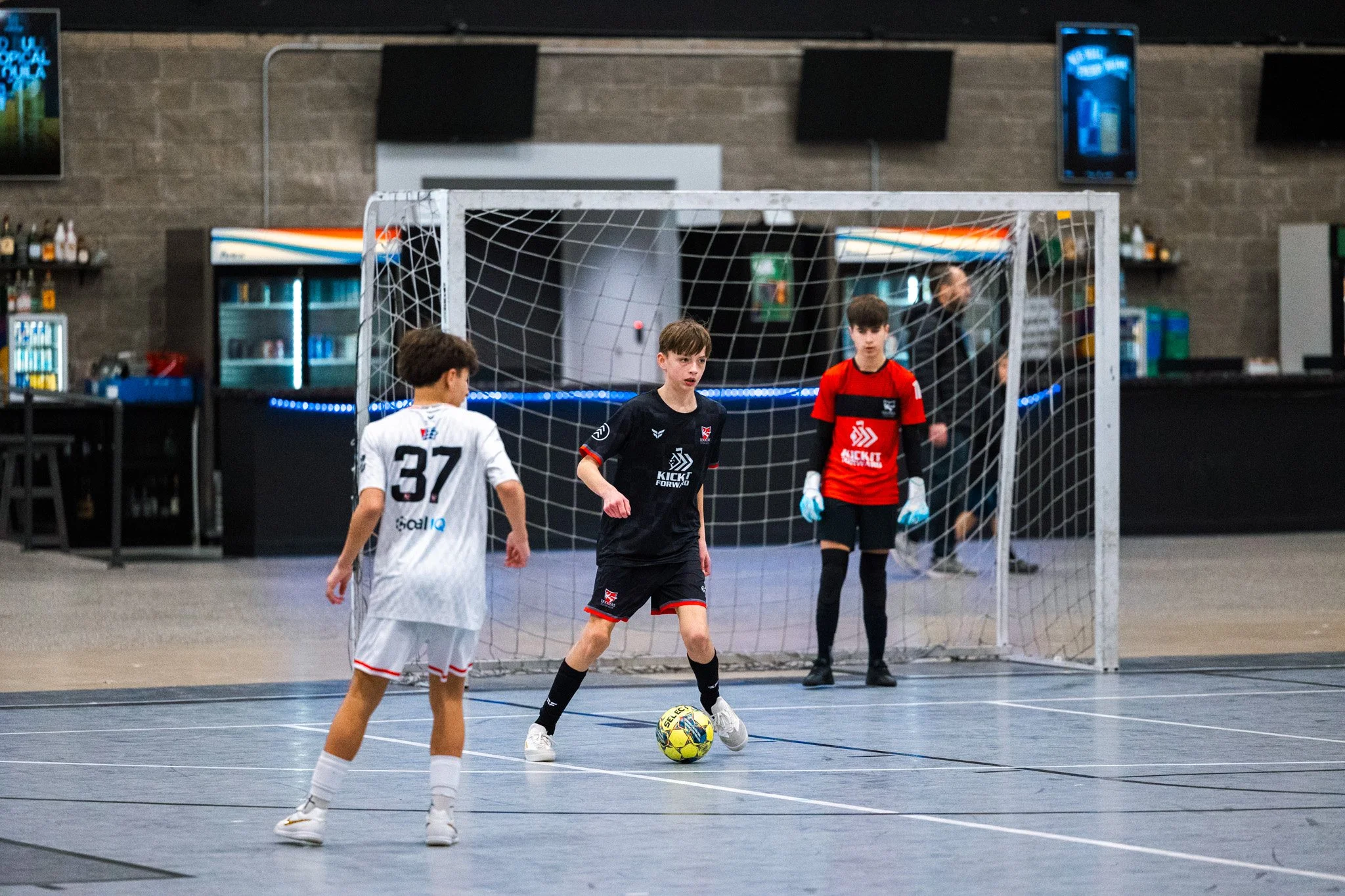 Young boys playing indoor soccer, one in a black jersey with a soccer ball, another in a white jersey, and a goalkeeper in a red jersey, on an indoor court with a netted goal.