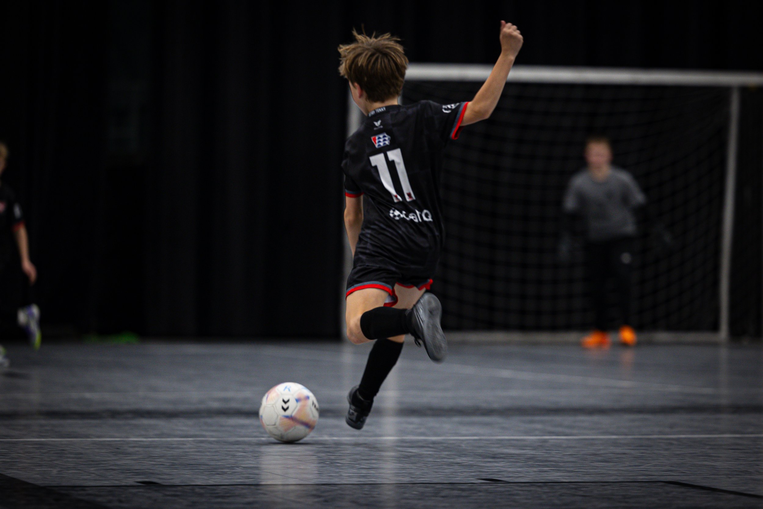 A young boy playing indoor soccer on a dark court, wearing a black sports uniform with the number 11, kicking a white soccer ball with black and blue markings, with a goal and a person in the background.
