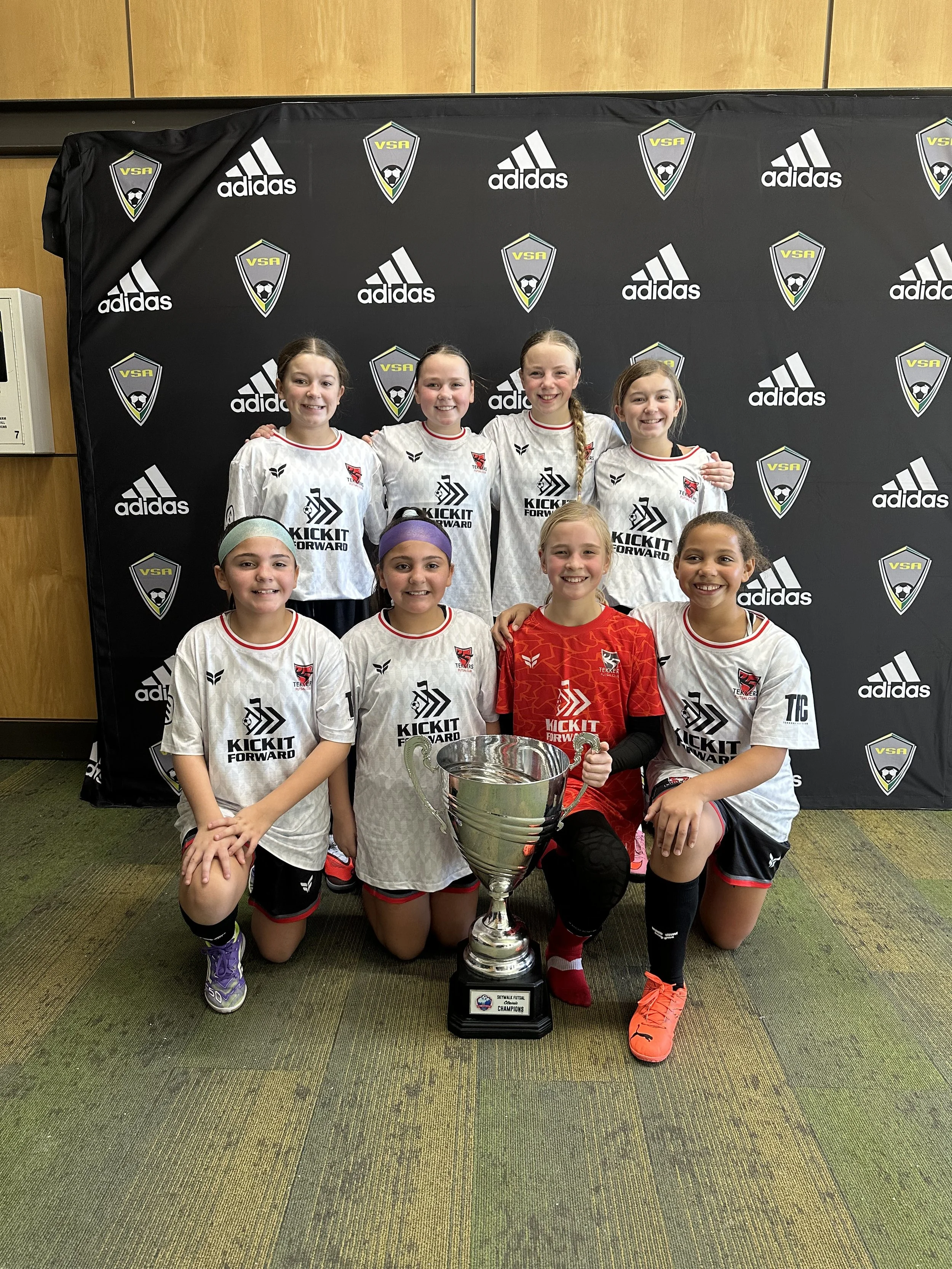 A girls' soccer team celebrating with a large silver trophy in front of a black backdrop decorated with Adidas and VSA logos. The team members are wearing white uniforms with red and black accents, and one girl in front is in a red goalkeeper jersey.