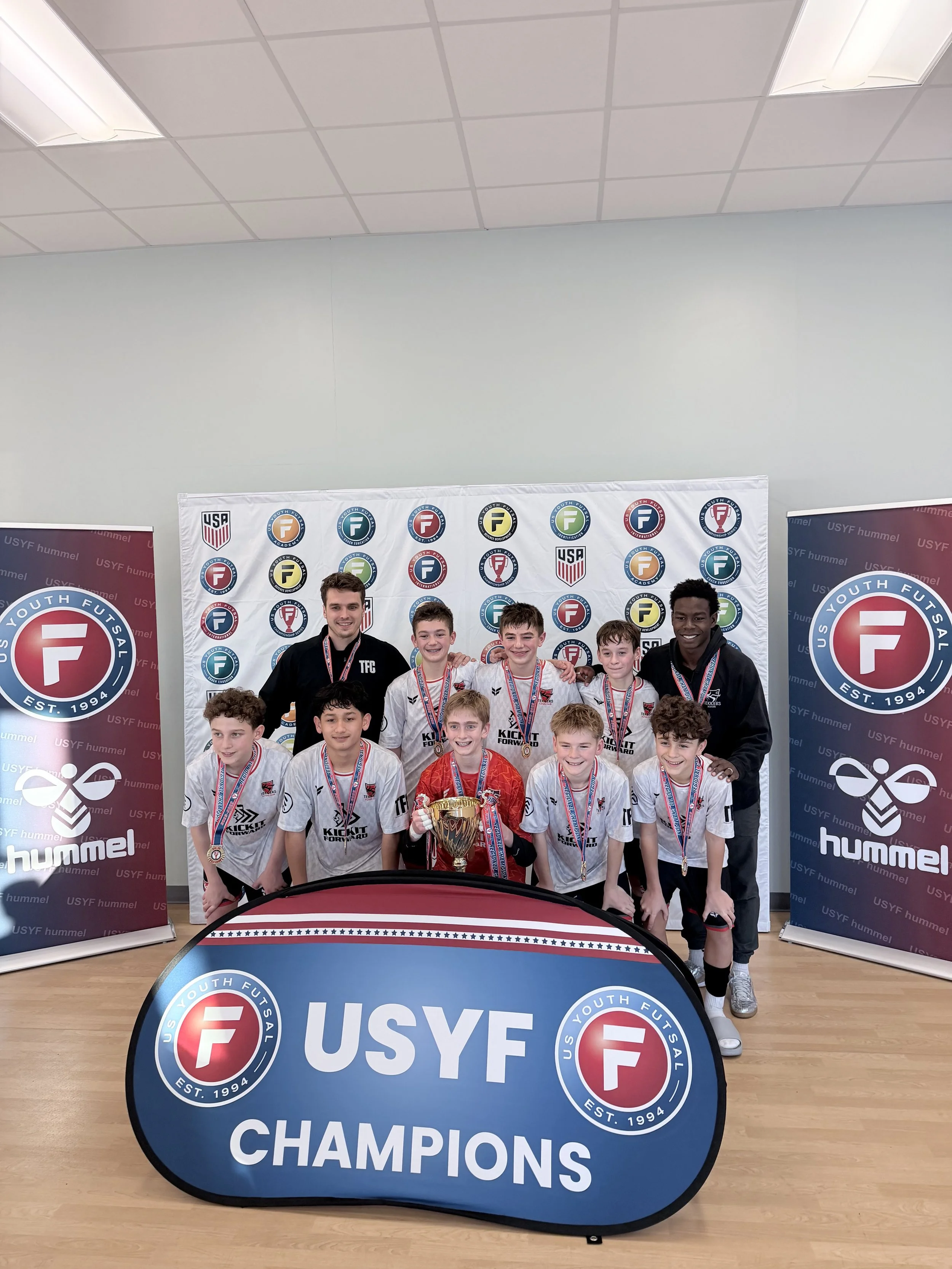 Group of young athletes and coaches posing with medals and a trophy at a youth futsal championship event, with banners displaying the US Youth Futsal logo and the words 'CHAMPIONS'.