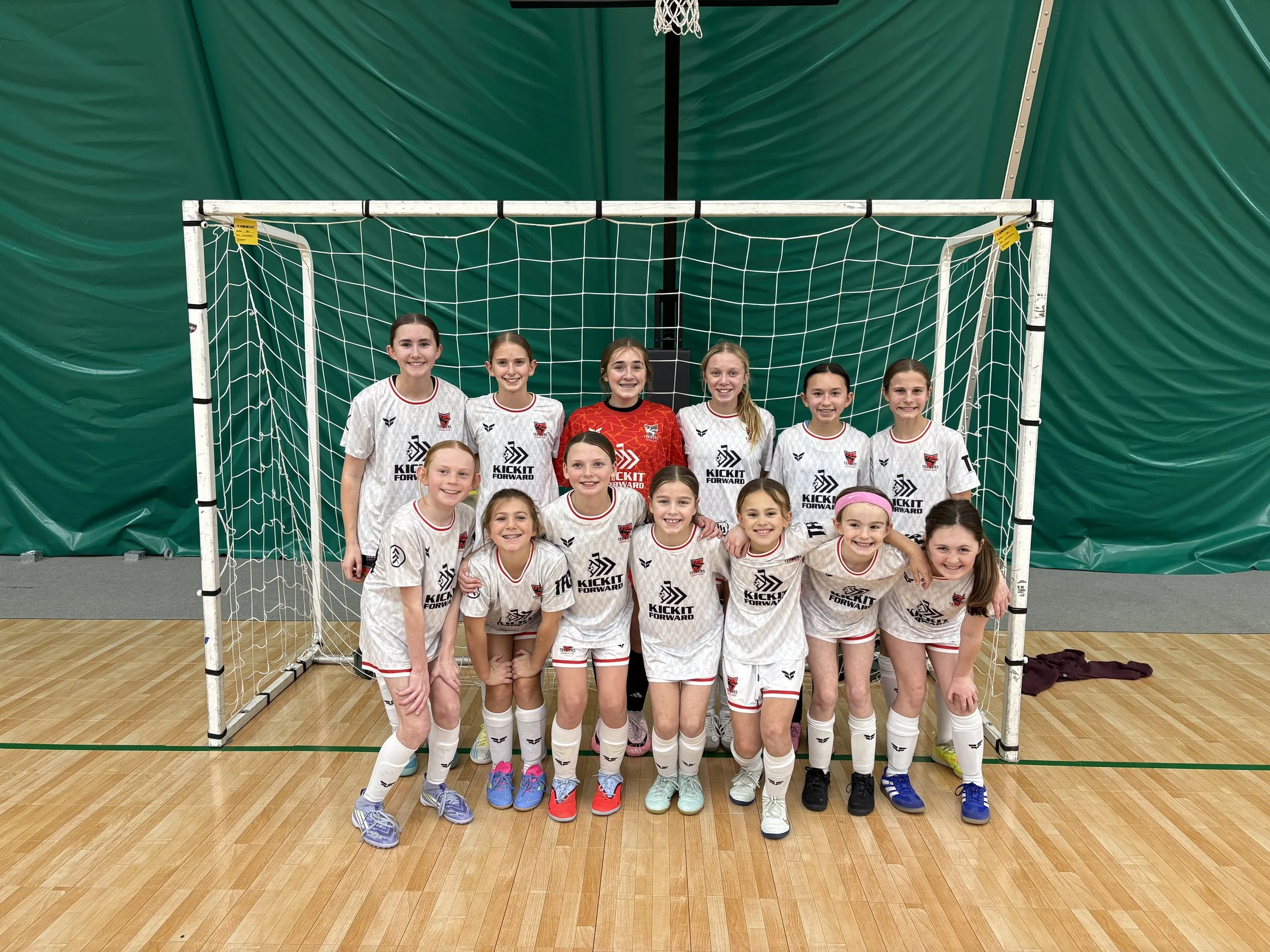 A group of young female soccer players in white and red uniforms standing together inside a sports hall, with a goal net behind them and a basketball hoop above.