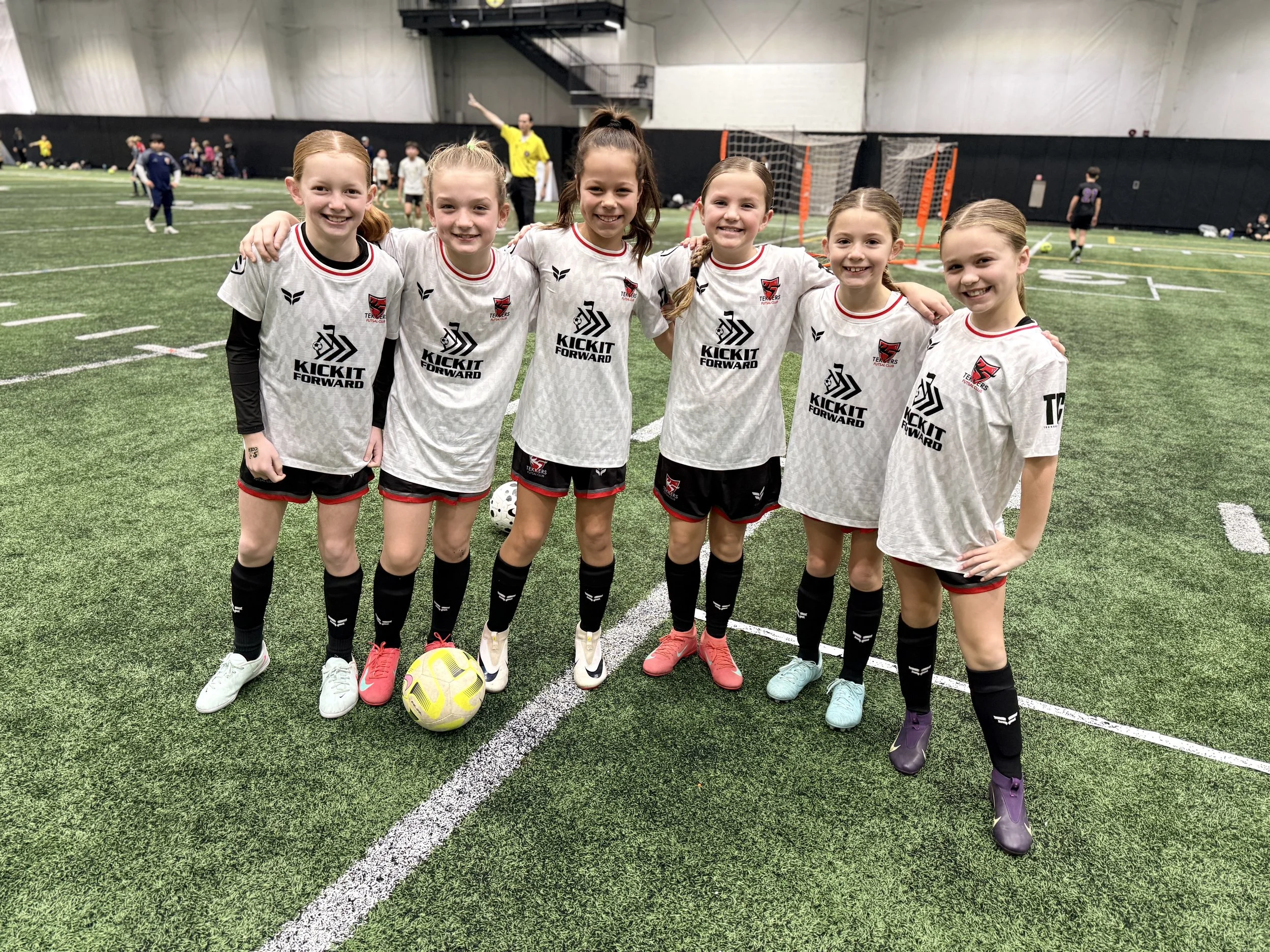 Six young girls in soccer uniforms with the text "Kick It Forward" and a logo on their jerseys, standing on an indoor soccer field. They are smiling with their arms around each other, and a yellow soccer ball is at their feet.