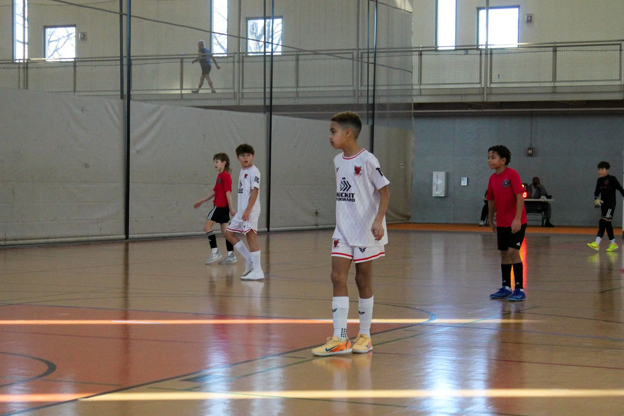 Young boys playing indoor soccer in a gymnasium, with some standing and others walking, one wearing a white uniform and others in red and black, while a woman in black goalie attire stands in the background near the wall.
