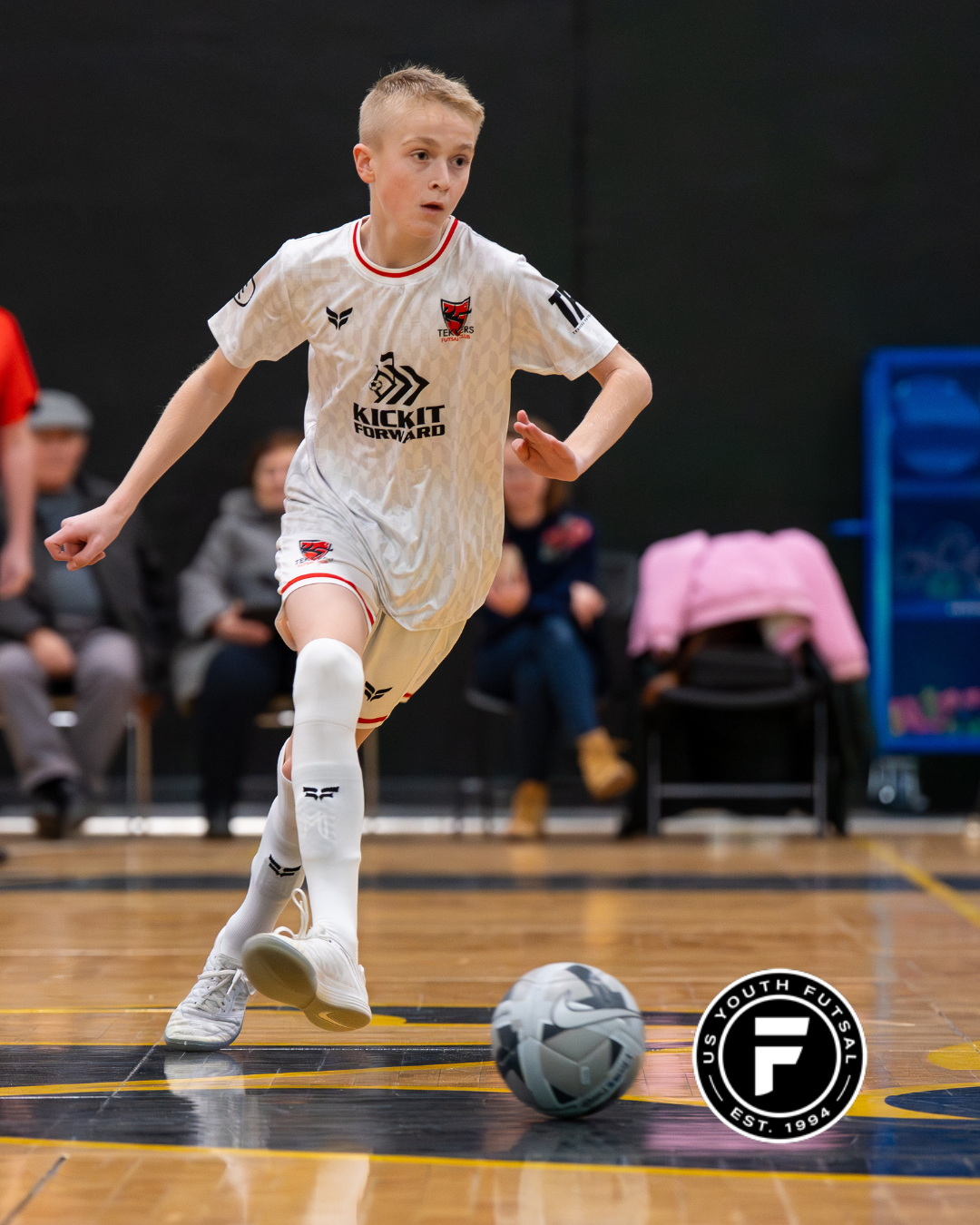 A young boy playing indoor soccer, wearing a white jersey with red accents, kicking a soccer ball on a wooden court with spectators in the background.