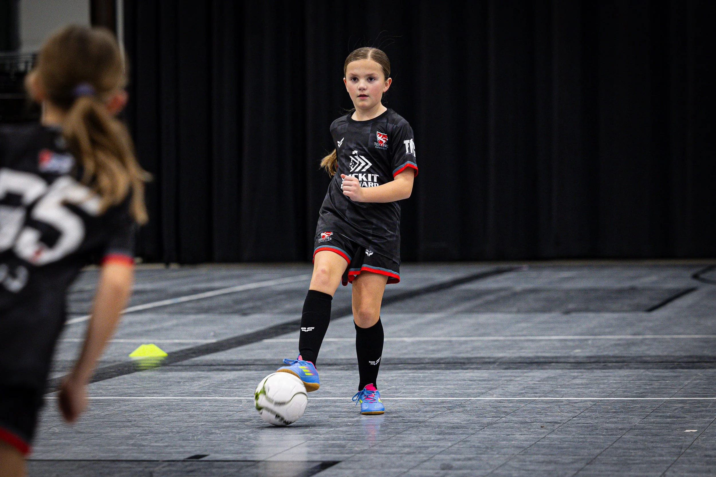 Young girl in sports uniform playing indoor soccer, preparing to kick a white soccer ball, with children in the background and a black curtain in the setting.